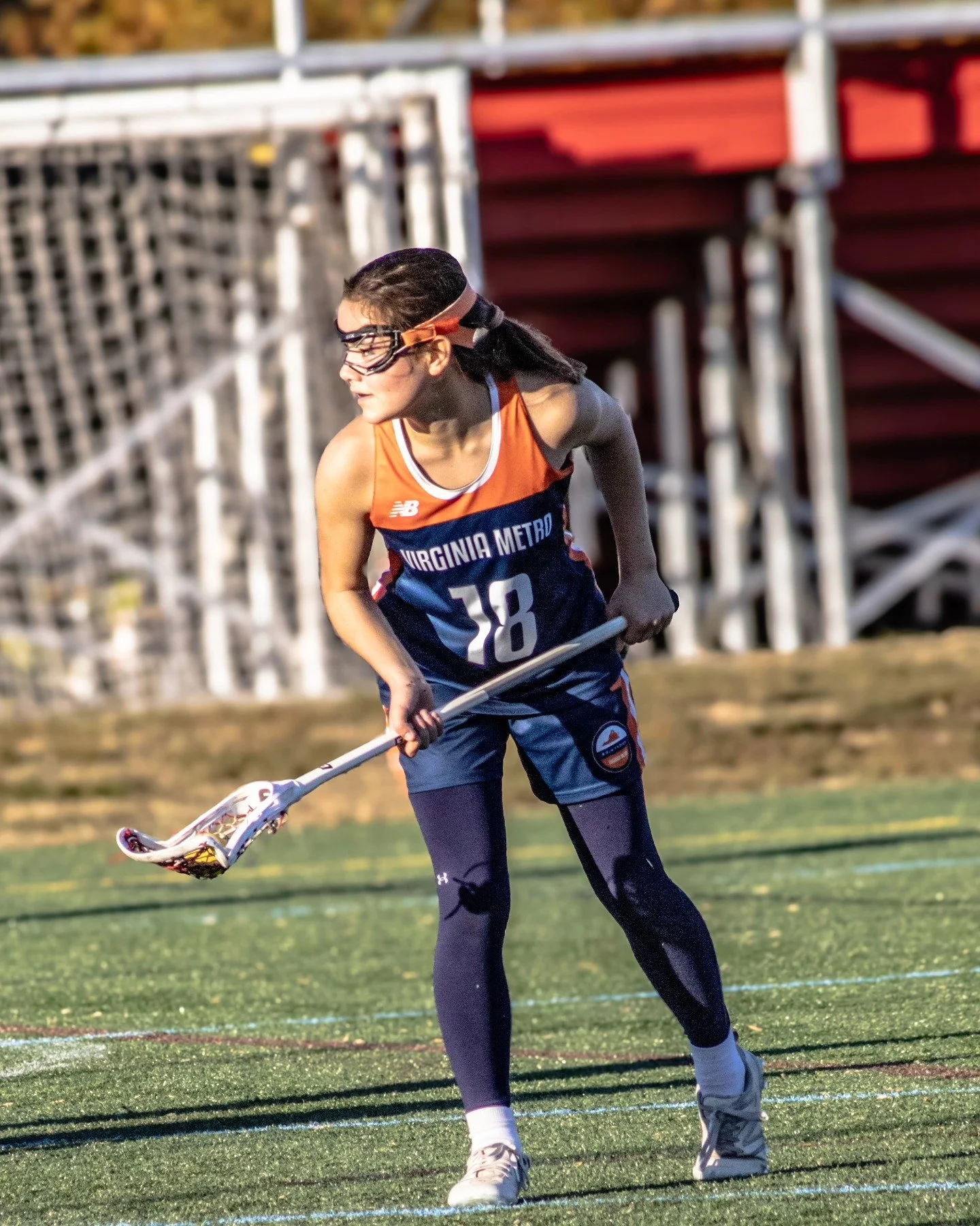 Sports photography Baltimore Maryland DMV. A female athlete with a lacrosse stick in hand on a grassy field during daytime, wearing sports gear including a Virginia Metro jersey, black leggings, sneakers, goggles, and a headband.