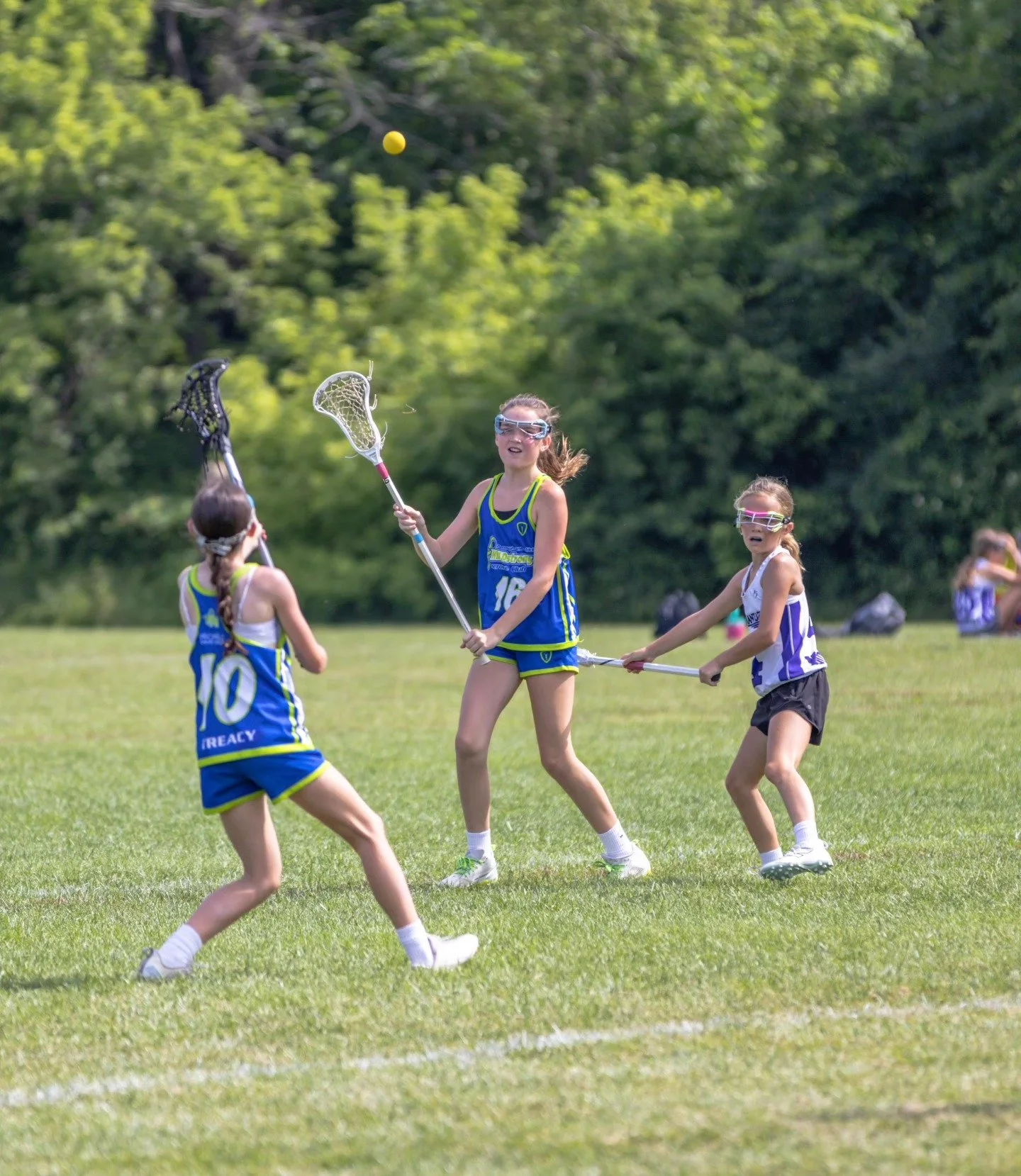 Sports photography Baltimore Maryland DMV. Three young girls playing lacrosse outdoors on a grassy field, wearing sports uniforms and protective eyewear, with a yellow ball in the air.