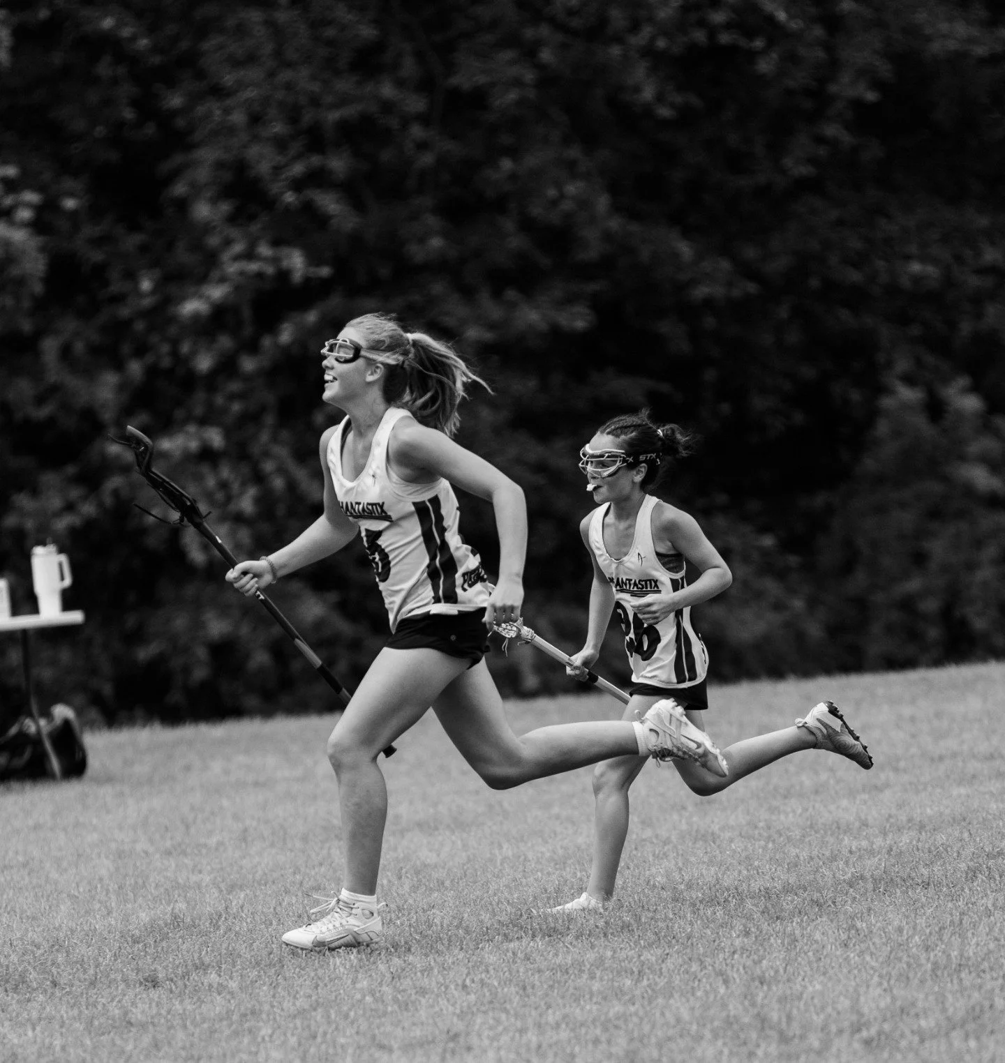 Sports photography Baltimore Maryland DMV. Two girls running on a grass field, wearing sports uniforms and sunglasses, holding poles, with trees in the background.