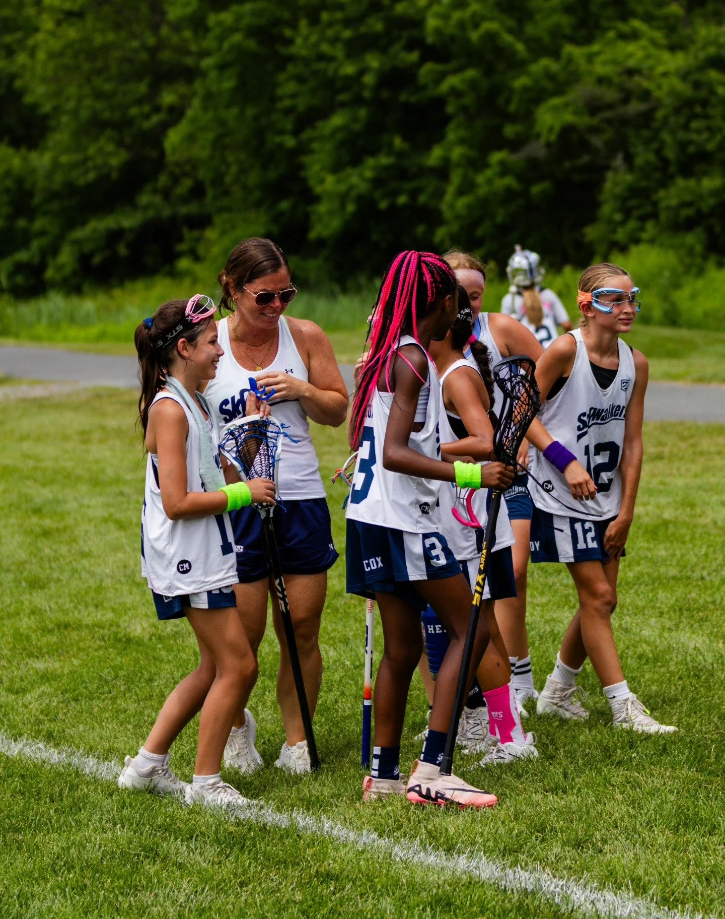 Sports photography Baltimore Maryland DMV. A group of young women and a coach standing on a grassy field, wearing athletic uniforms and holding lacrosse sticks, engaged in a team discussion.