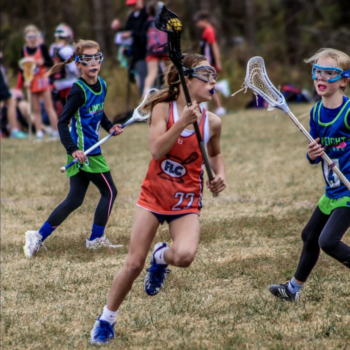 Sports photography Baltimore Maryland DMV. Youth girls playing lacrosse outdoors, running on grass field, wearing uniforms, goggles, and holding lacrosse sticks.