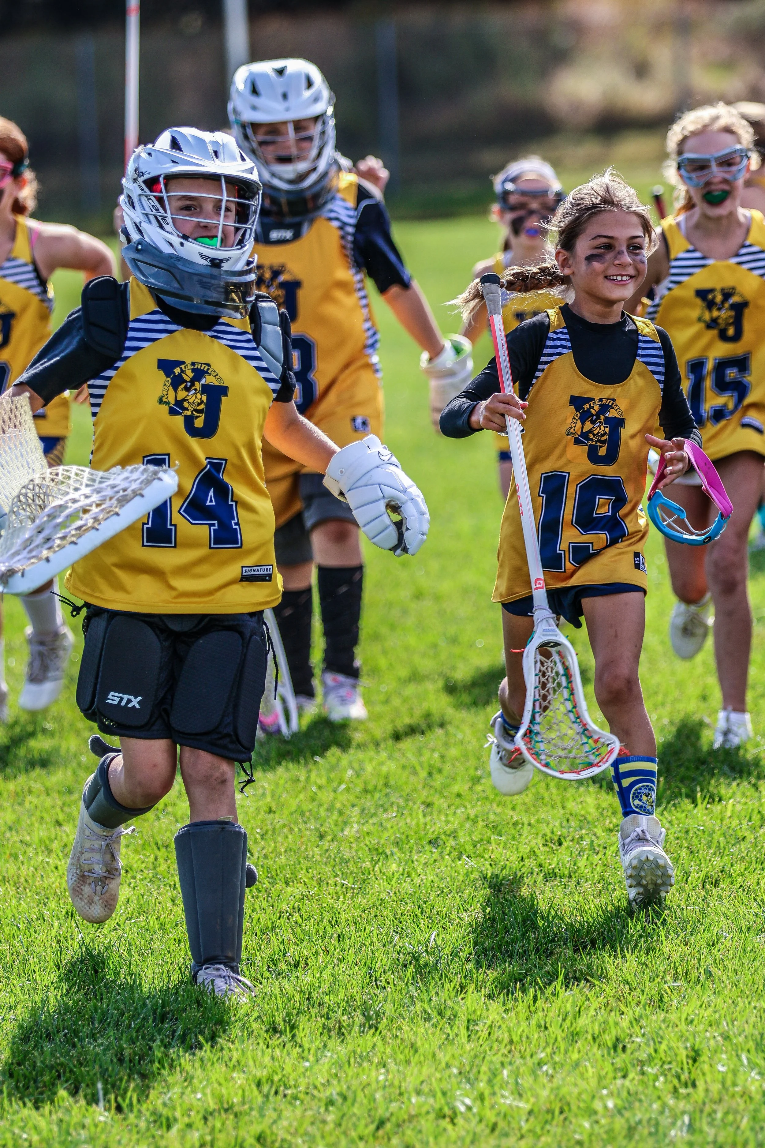 Sports photography Baltimore Maryland DMV. Young girls and boys in yellow sports jerseys playing lacrosse on a grassy field during daytime, some wearing helmets and protective gear, smiling and running.