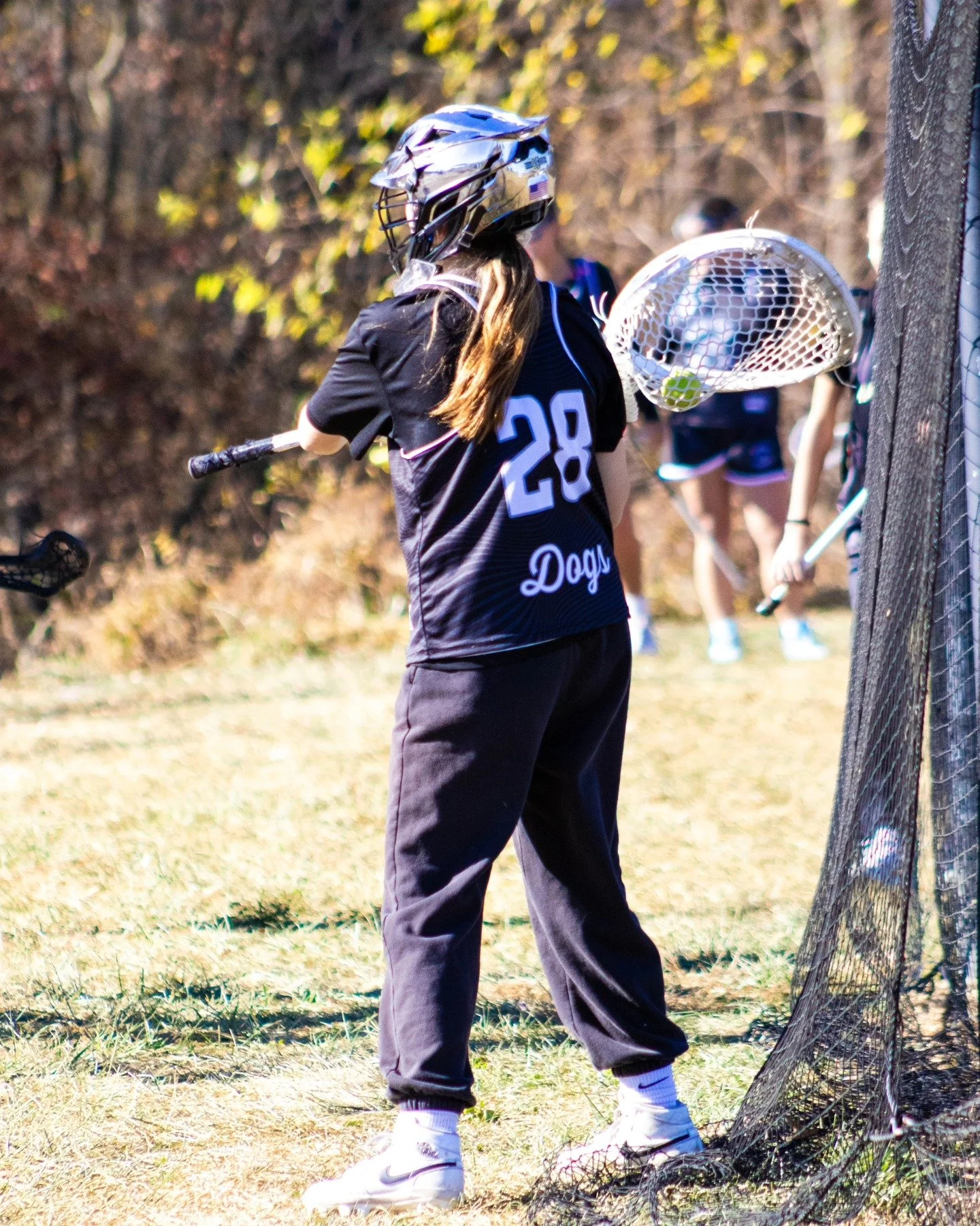 Sports photography Baltimore Maryland DMV. A girl wearing a sporty black jersey with the number 28 and the word 'Dogs' written below it, a helmet, and holding a lacrosse stick, is playing lacrosse outdoors near a net with a ball inside the net.