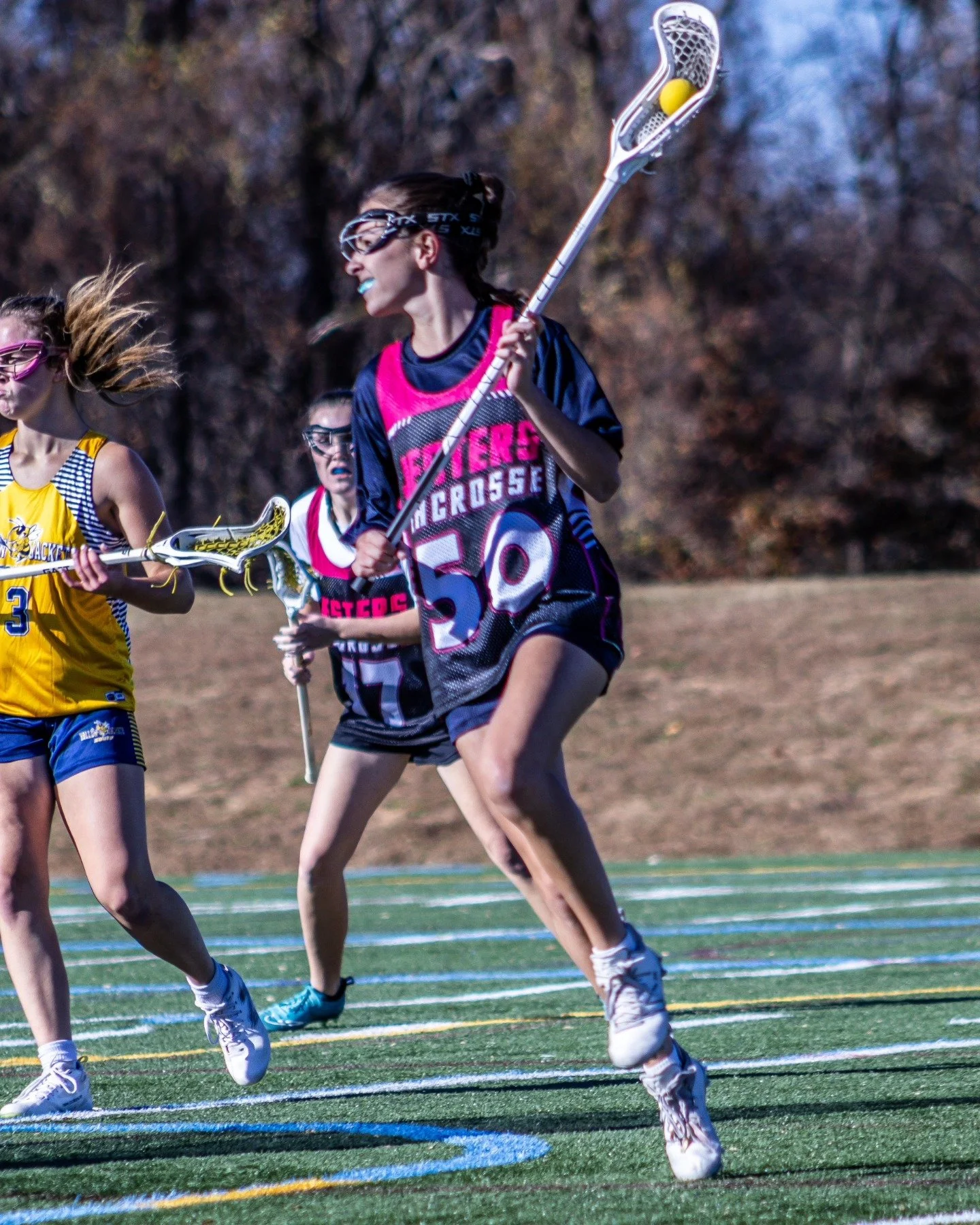 Sports photography Baltimore Maryland DMV. Lacrosse players running on a field during a game, with one player holding a lacrosse stick with a yellow ball, wearing sports gear, and wearing glasses or protective eyewear.