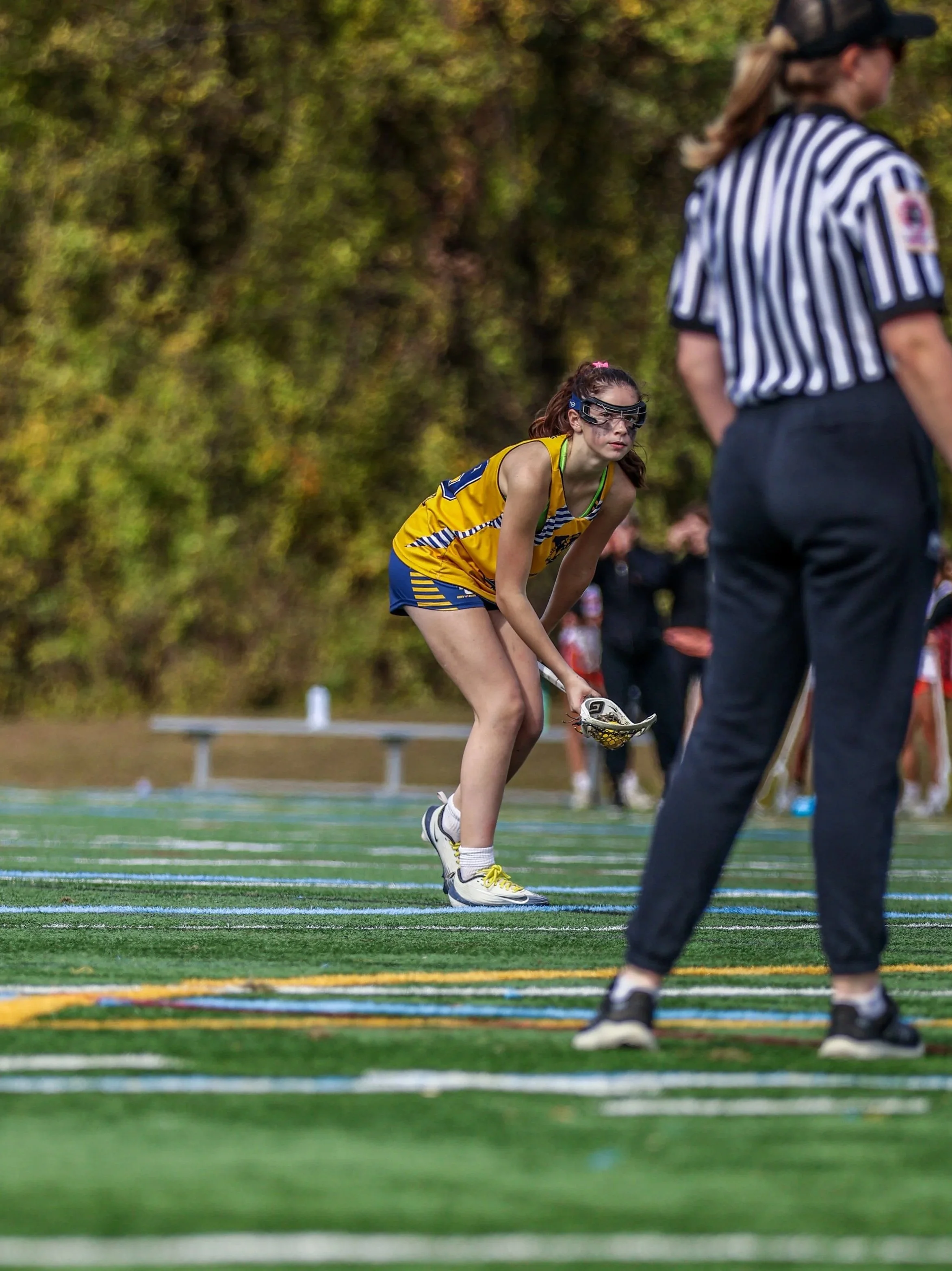 Sports photography Baltimore Maryland DMV. A young female lacrosse player in a yellow and blue uniform is preparing to catch or hold her lacrosse stick on a field during a game or practice, with officials and other players visible in the background.