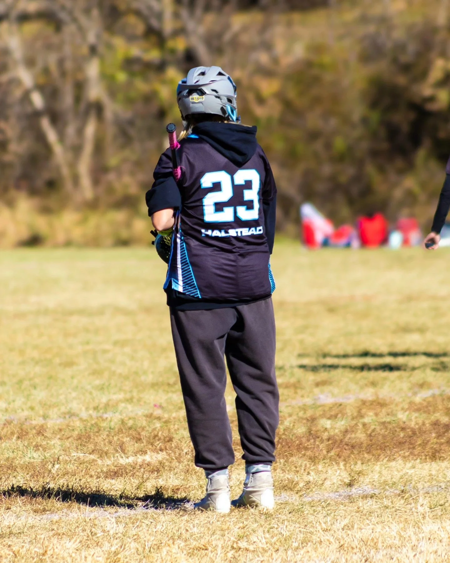 Sports photography Baltimore Maryland DMV. Person wearing a gray helmet, black Nike jacket with the number 23 and the word 'HALSTEAD' on the back, gray pants, and white shoes, standing on a grassy field during daytime.