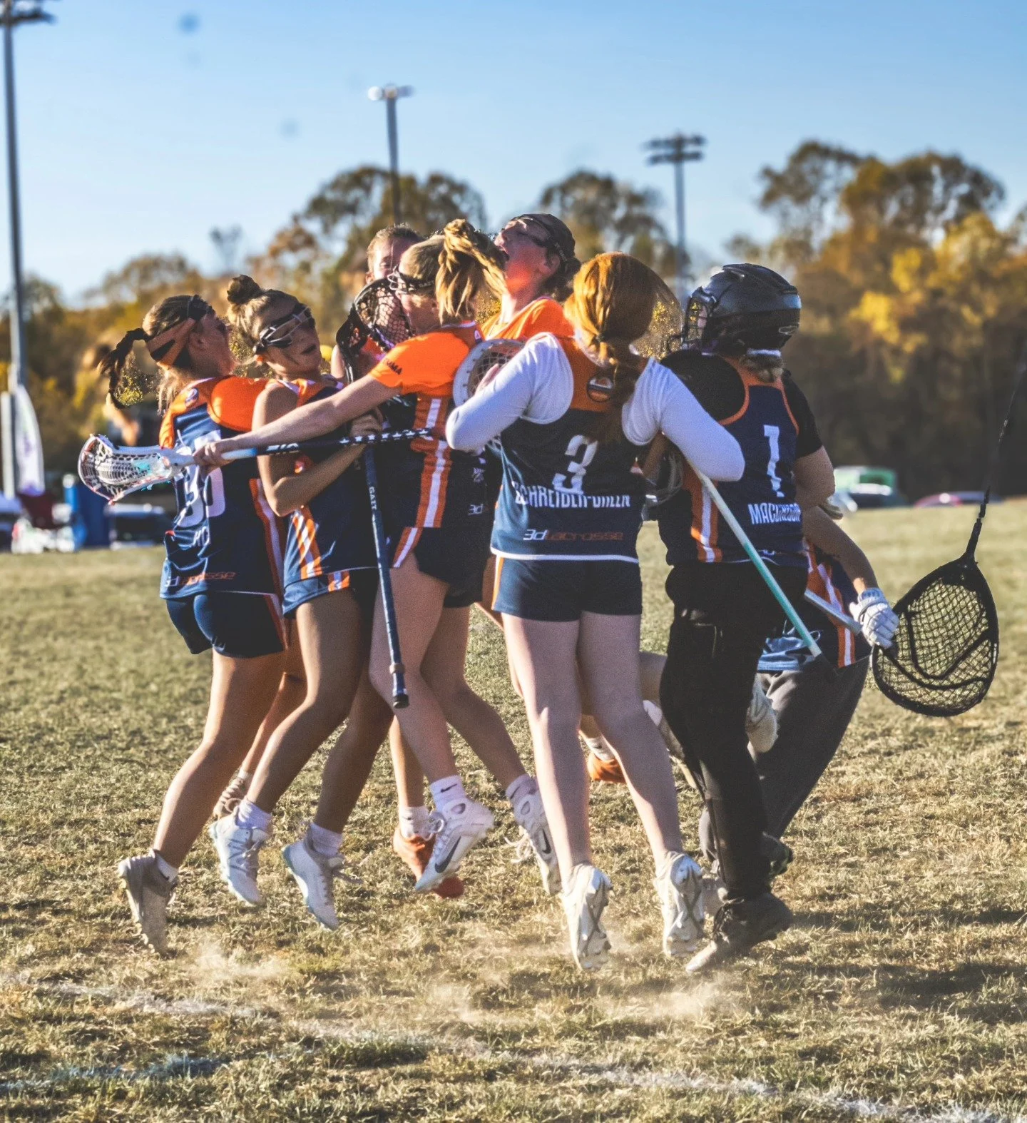 Sports photography Baltimore Maryland DMV. Girls' lacrosse team celebrating together on a grass field during daytime, some players are hugging and some are holding lacrosse sticks, with trees and a clear sky in the background.