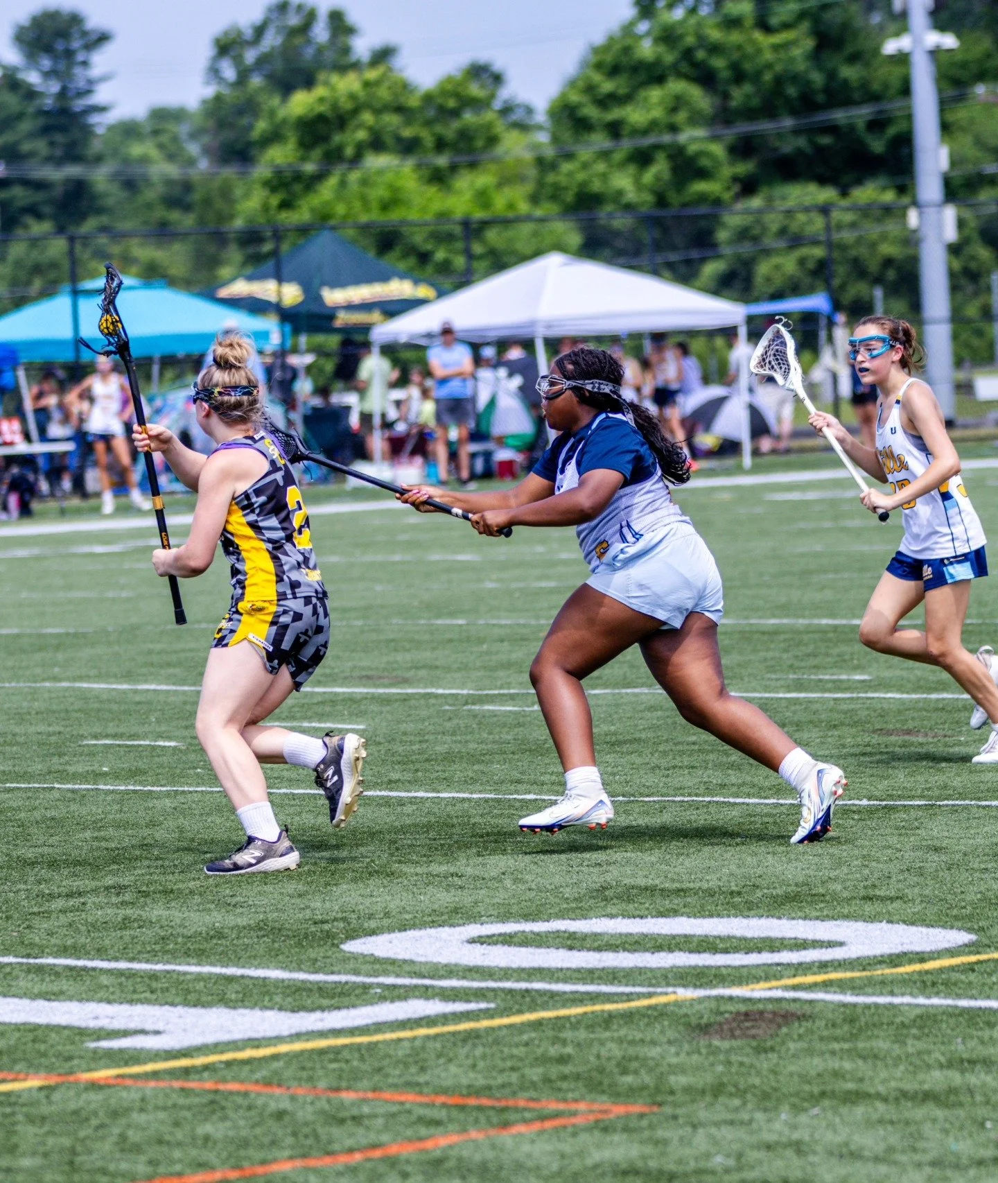 Sports photography Baltimore Maryland DMV. Three young female lacrosse players running on a green field during a game, wearing athletic gear and goggles, with tents and spectators in the background.