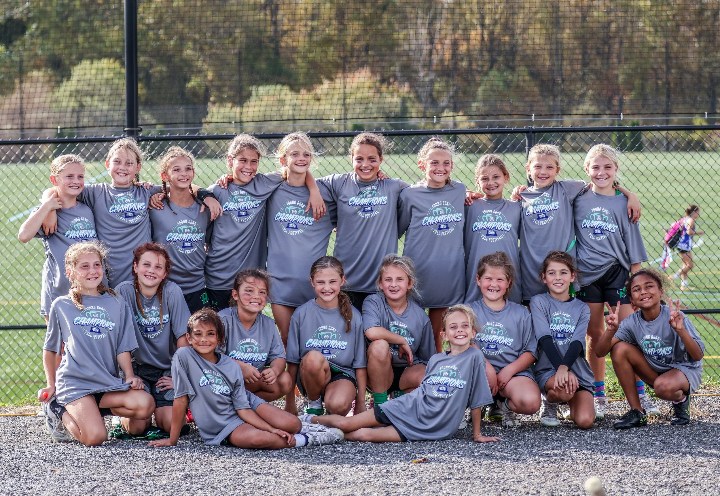Sports photography Baltimore Maryland DMV. A group of young girls in matching gray T-shirts with 'Champions' printed on them, posing for a team photo on a sports field with a school fence and trees in the background.