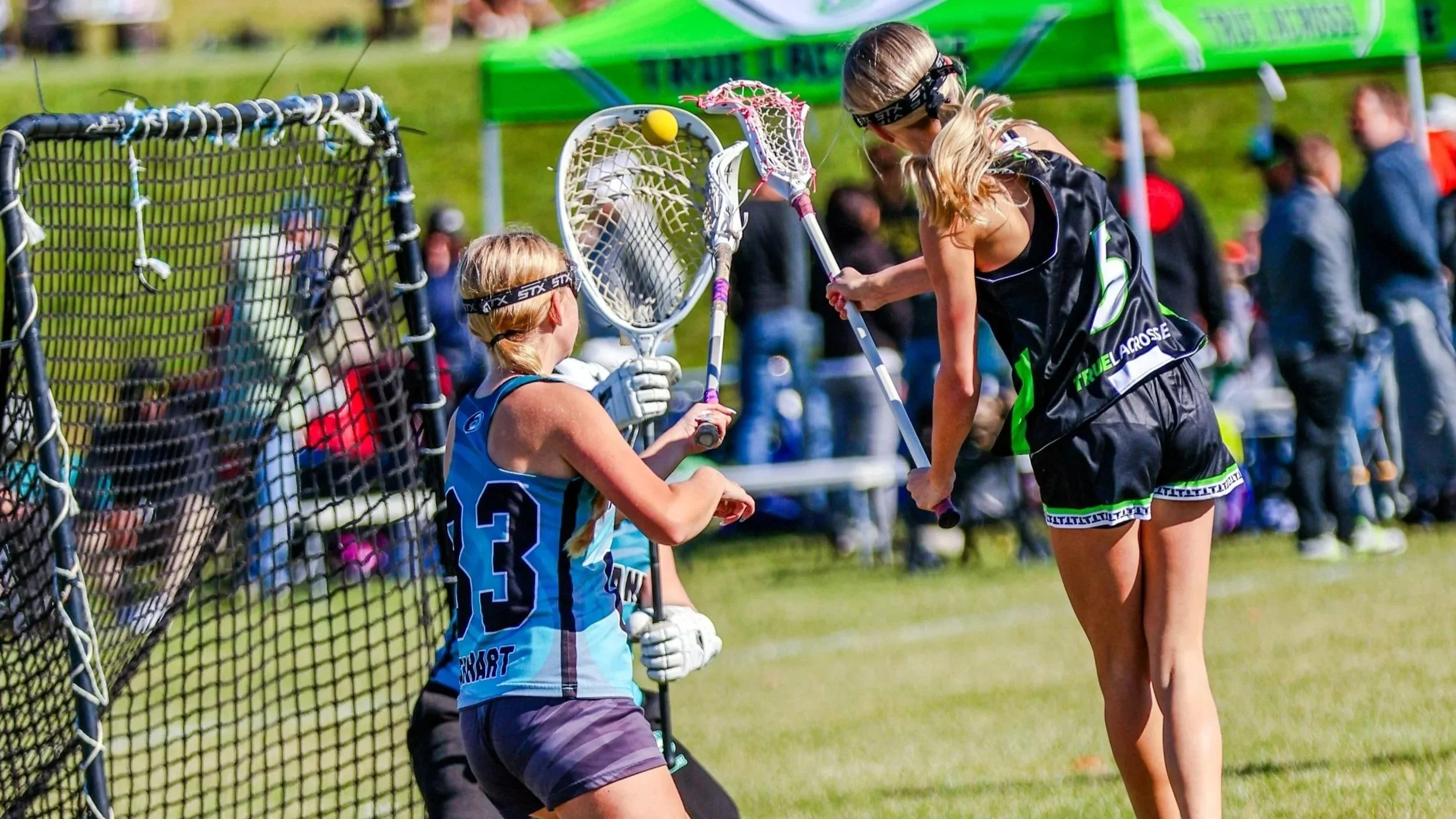 Sports photography Baltimore Maryland DMV. Two female field hockey players, one sitting and one standing, exchanging a ball near a goalpost during a game on a grassy field, with spectators in the background.