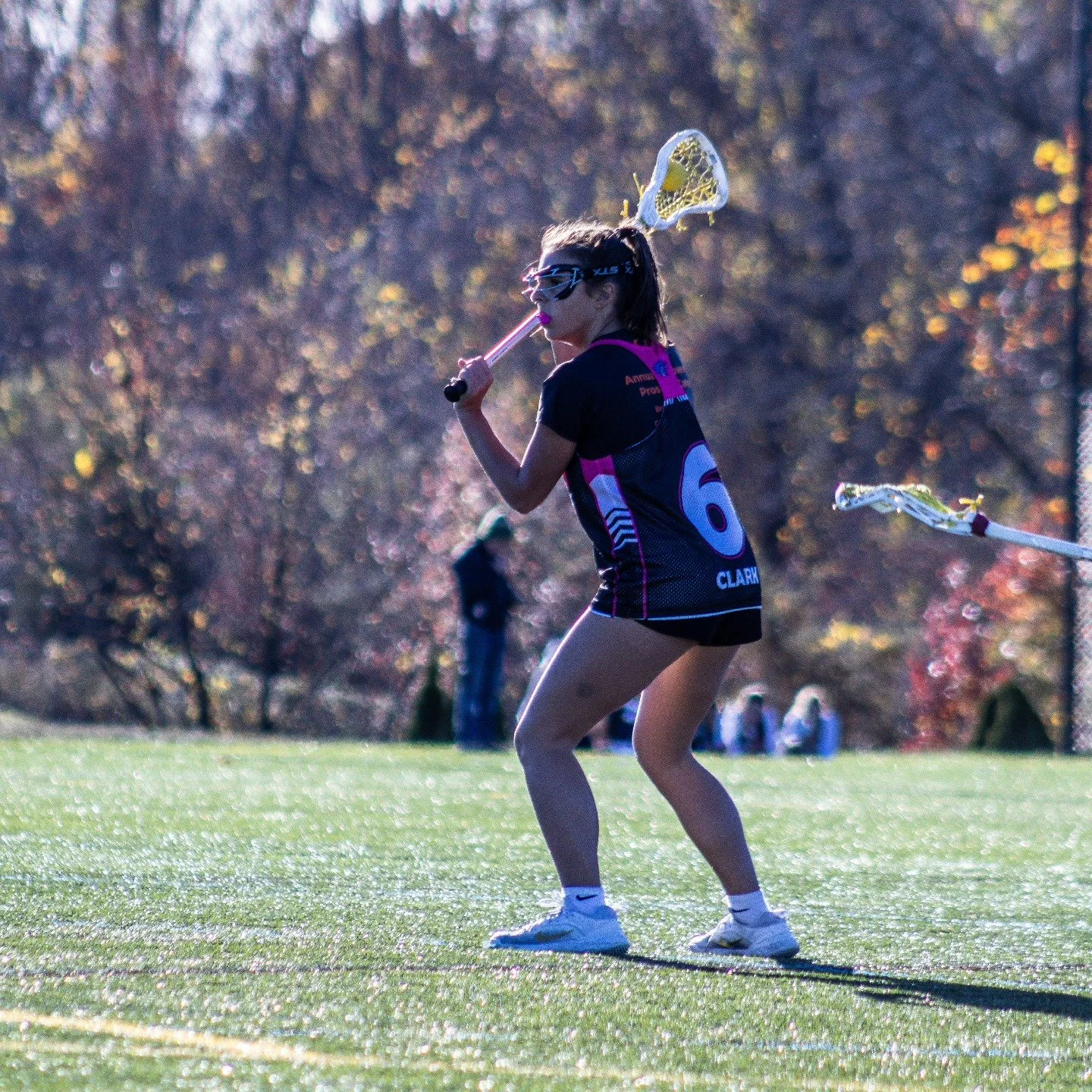 Sports photography Baltimore Maryland DMV. A young woman playing lacrosse on a grassy field during daytime, wearing protective goggles and a jersey with the number 6, holding a lacrosse stick with a net, with autumn trees in the background.