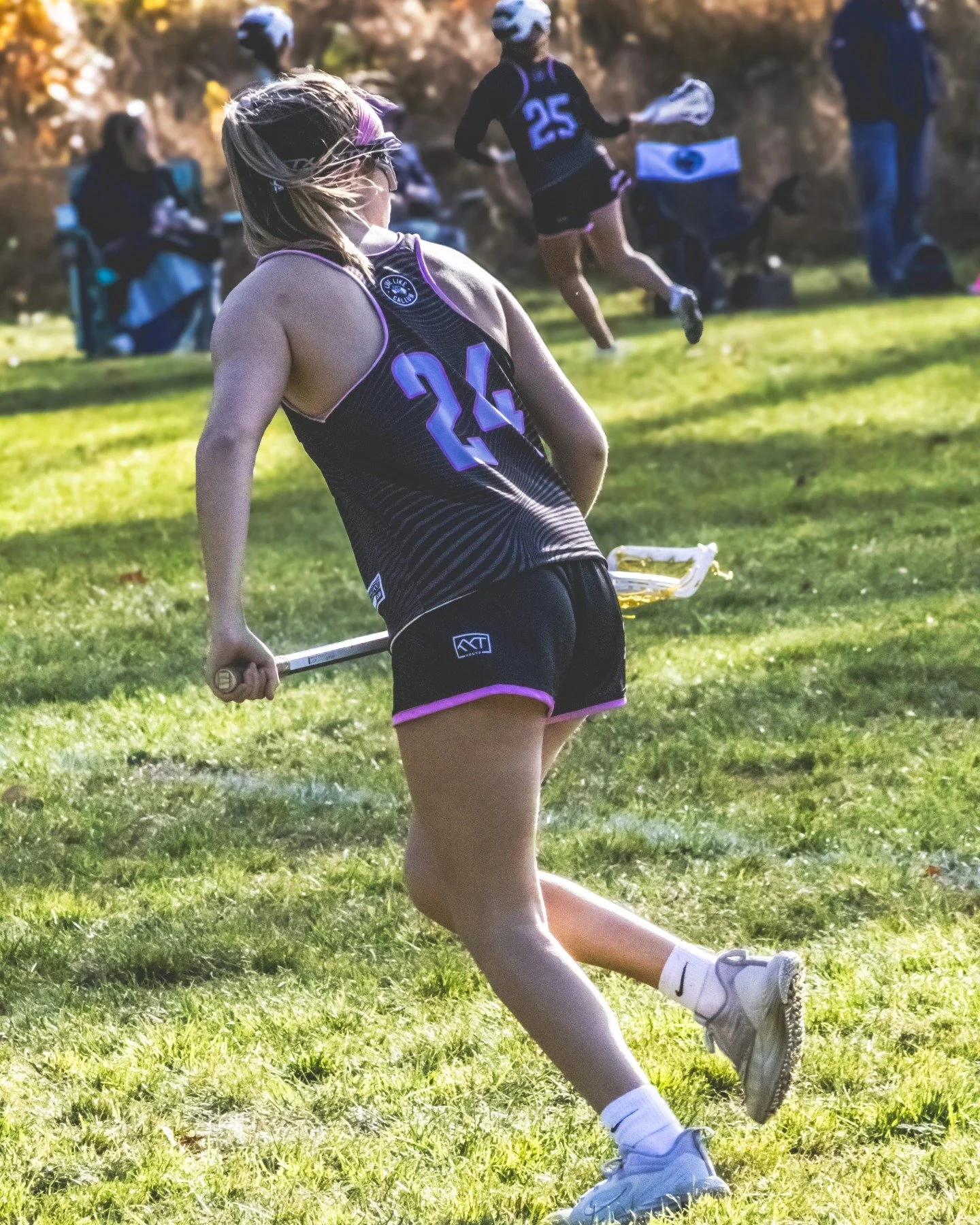 Sports photography Baltimore Maryland DMV. A woman wearing a black and purple sports uniform, holding a lacrosse stick, standing on a grassy field during a game or practice. There are other people and lacrosse equipment in the background.