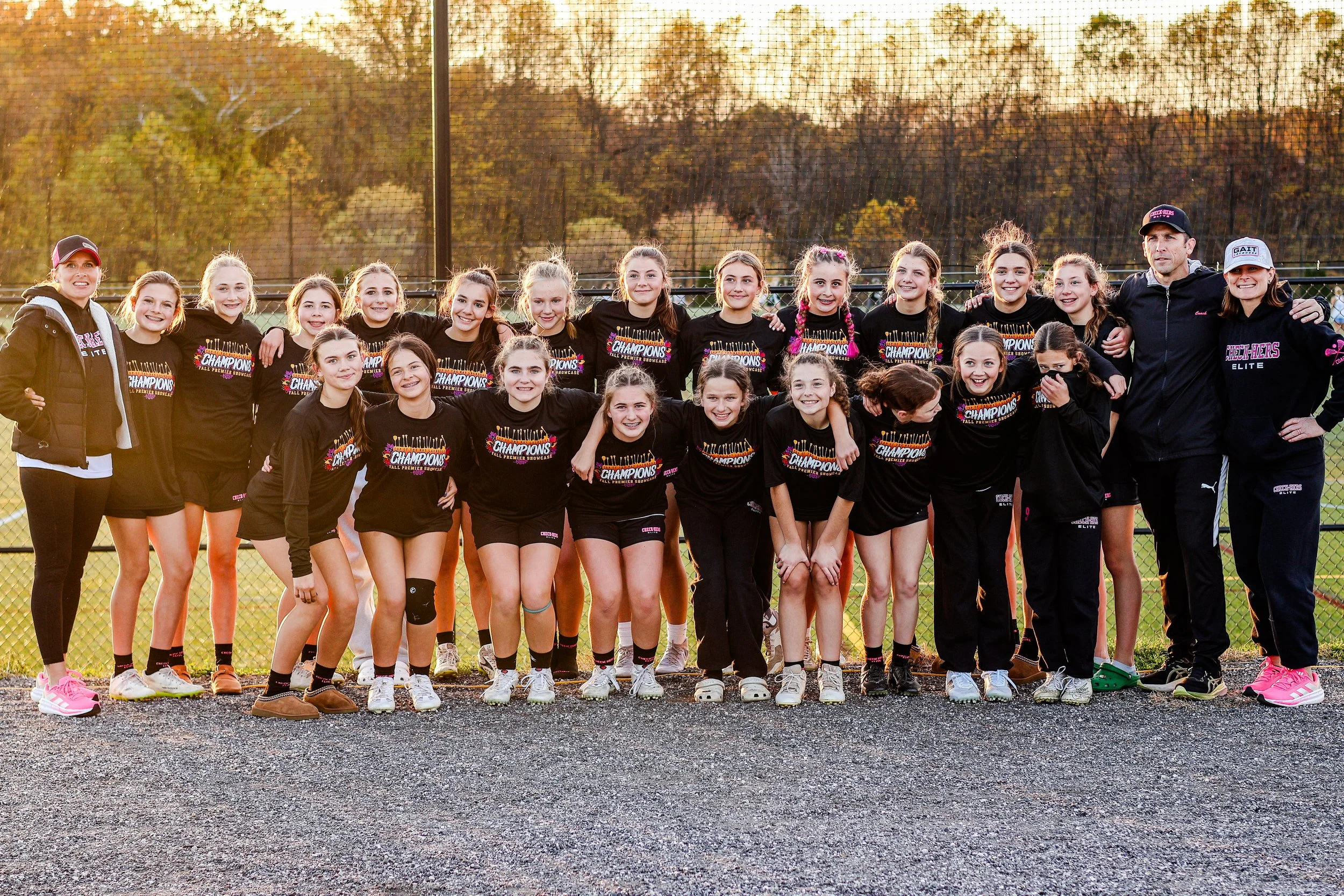 Sports photography Baltimore Maryland DMV. A group of young female athletes and their coaches pose together outdoors on a tennis court,"Champions." The girls and coaches are smiling with their arms around each other