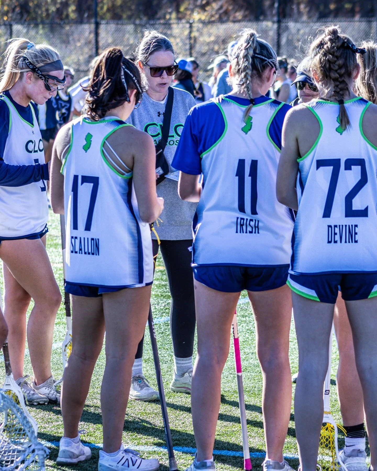 Sports photography Baltimore Maryland DMV. Women’s lacrosse team in huddle for coaching during daytime game on field, wearing white and blue jerseys with green accents and numbers, holding lacrosse sticks, with coach speaking.