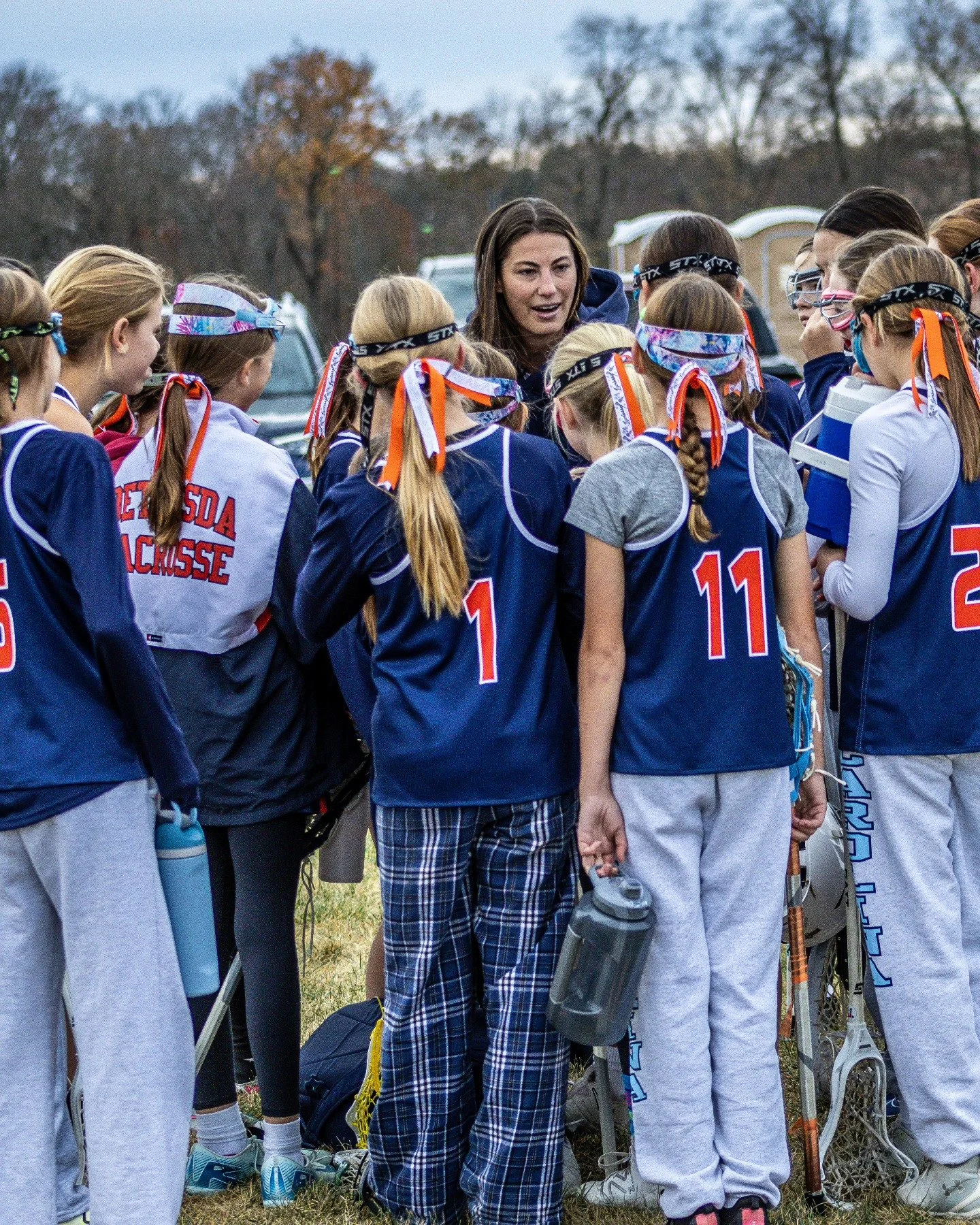 Sports photography Baltimore Maryland DMV. A female coach or team leader speaks to a group of young female lacrosse players who are gathered outdoors on a field, holding lacrosse sticks, during daytime.