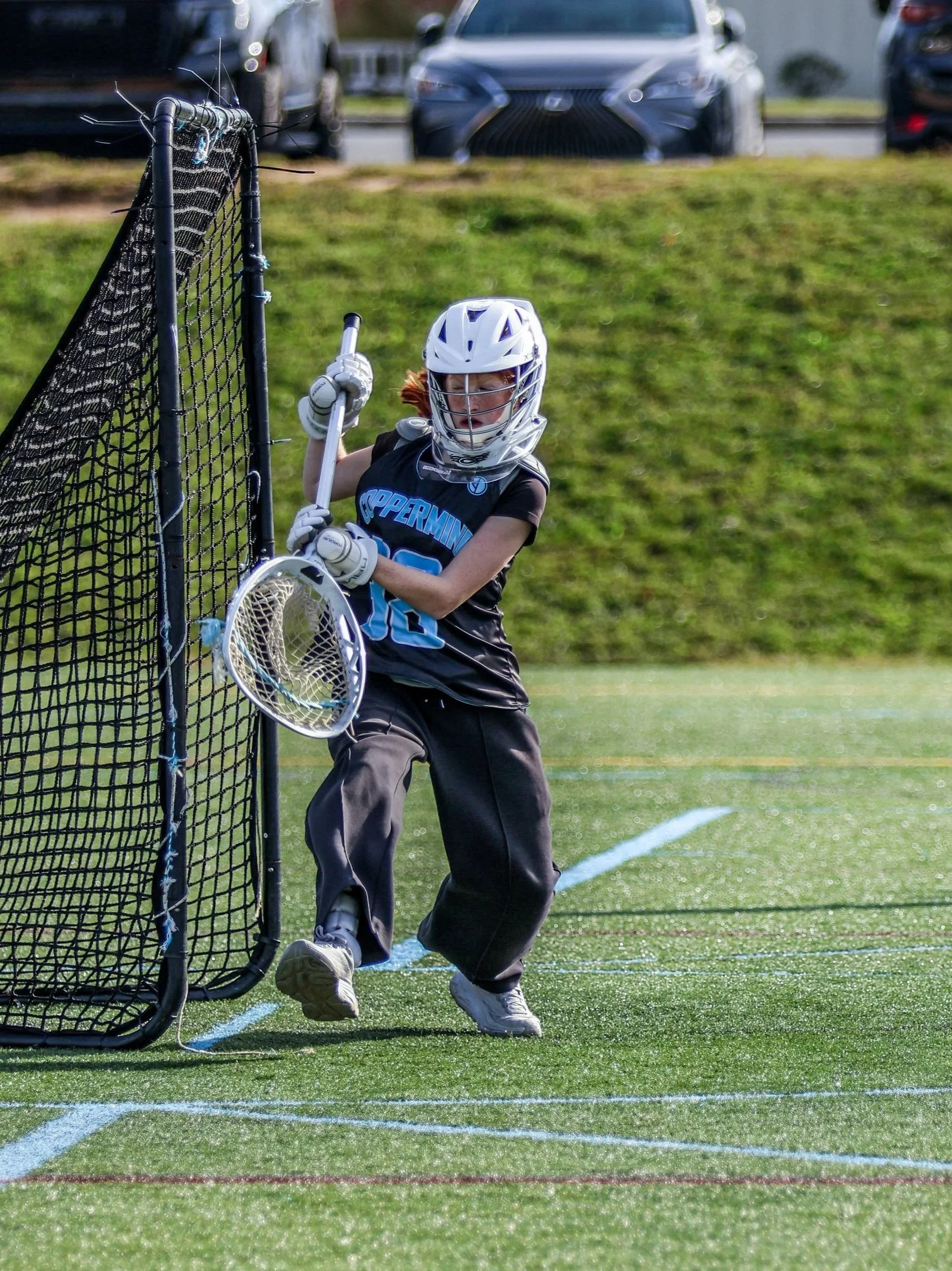 Sports photography Baltimore Maryland DMV. A young lacrosse player wearing a helmet and black jersey is running on a green field near a lacrosse goal, with parked cars visible in the background.