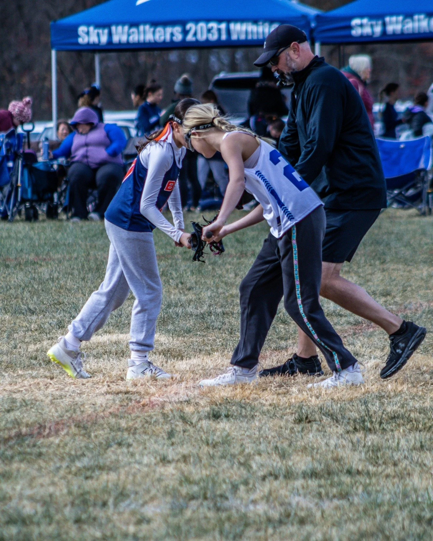 Sports photography Baltimore Maryland DMV. Two runners with a man helping one pick up a shoe on a grassy field during a race or event, with a crowd and tents in the background.