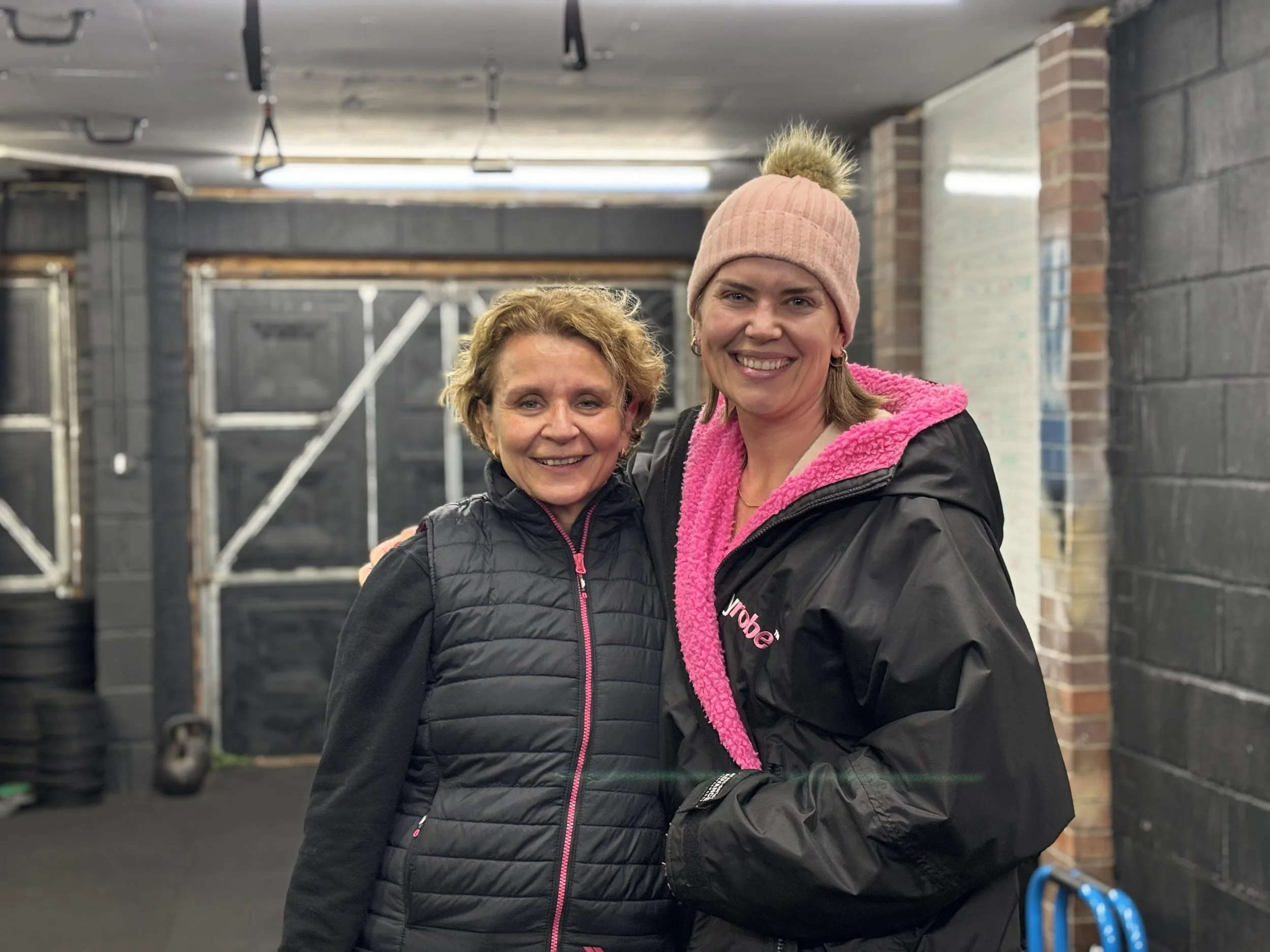 Two smiling women standing together inside of a gym, with one woman wearing a pink beanie and a black jacket with pink accents, and the other woman wearing a black vest with pink zipper and lettering, and a black jacket with a pink hoodie lining.