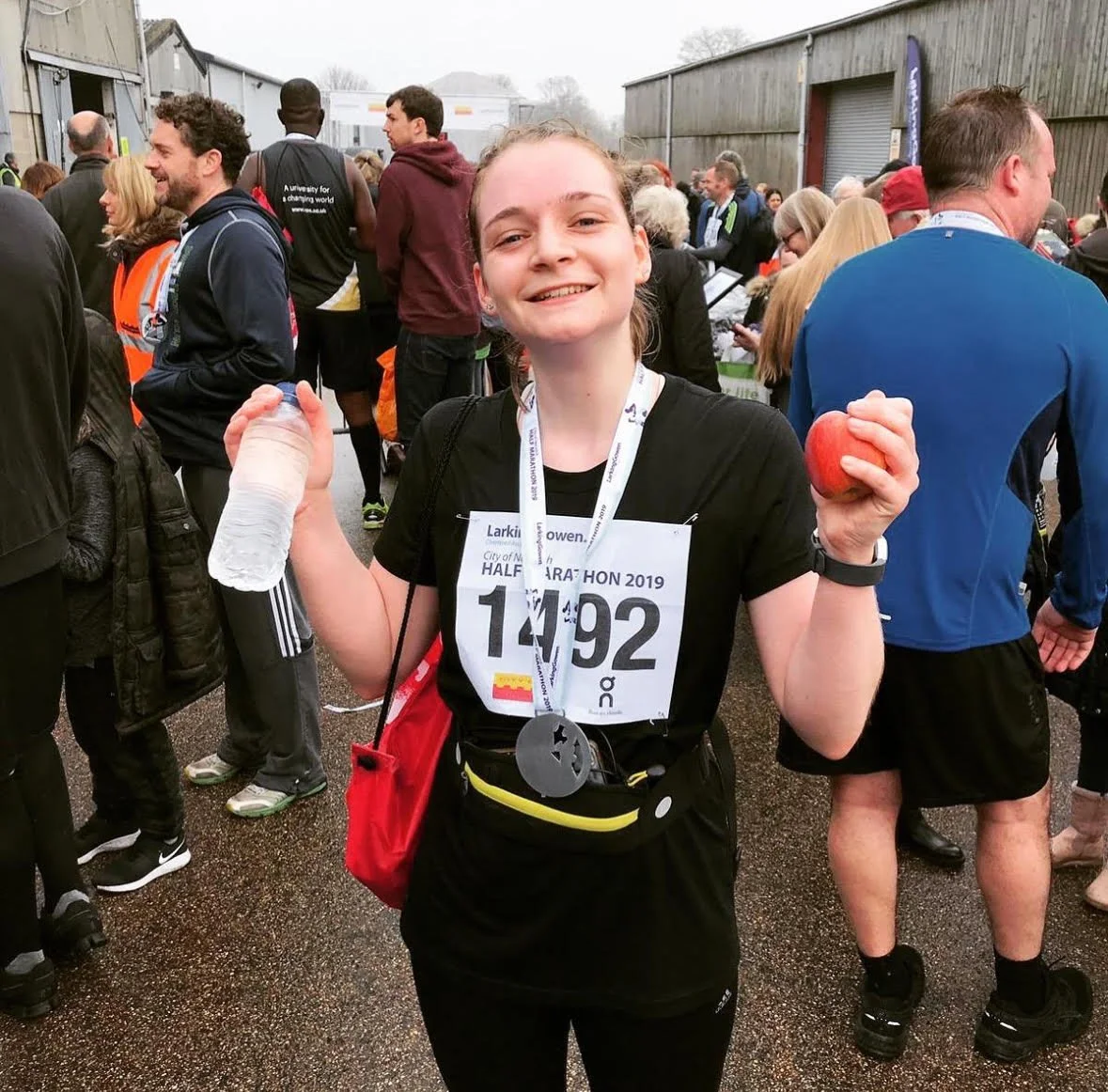 A young woman smiling at the finish of a half marathon, holding a water bottle and an apple, wearing a race bib number 1492, surrounded by other runners and spectators.