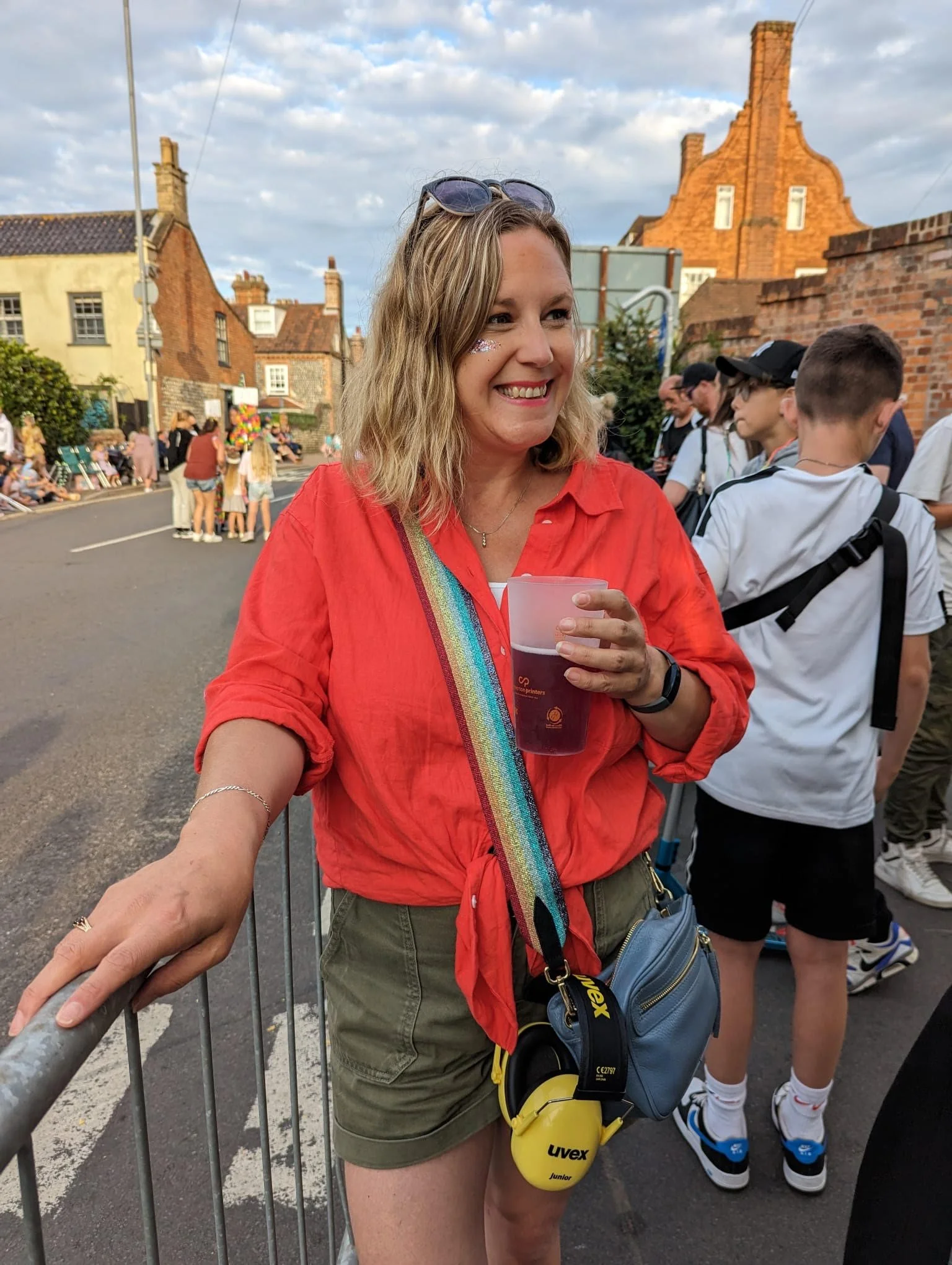 Smiling woman in red shirt holding a drink, standing outdoors at a festival or event, with people and historic buildings in the background.