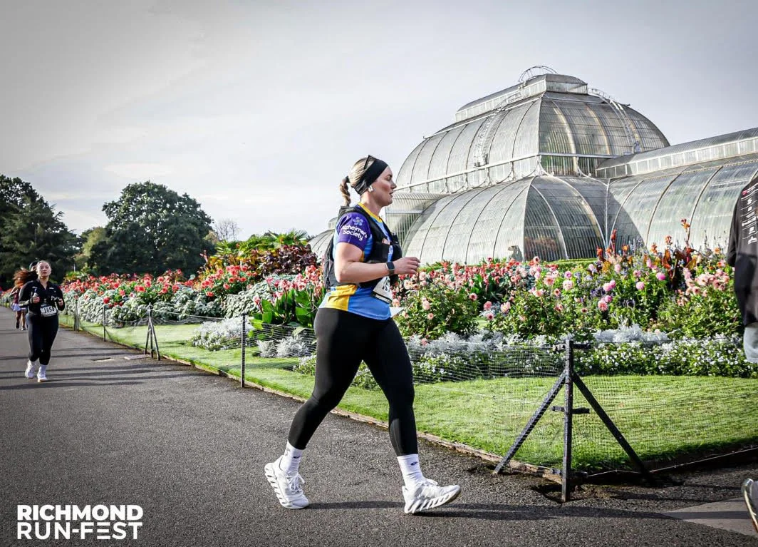 A woman running in a race next to a greenhouse with colorful flowers, with others behind her and the Richmond Run-Fest logo in the bottom left corner.