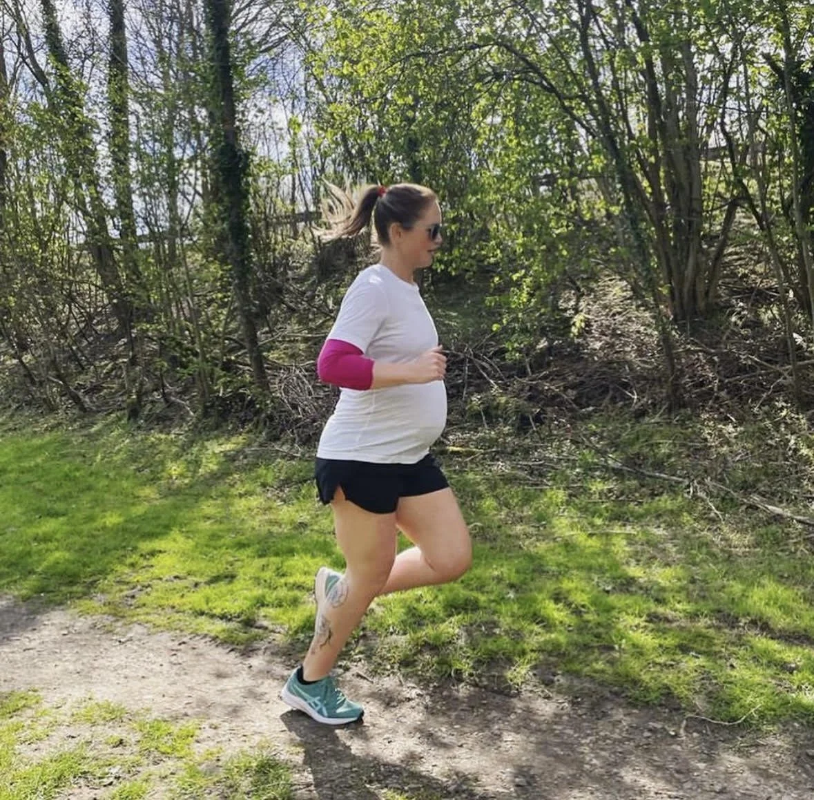 A pregnant woman running outdoors on a trail through a wooded area during daytime.