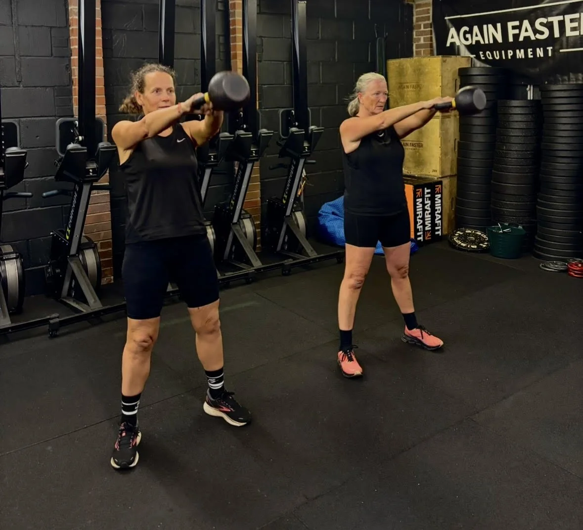 Two women performing a kettlebell shoulder press in a gym, with workout equipment and a banner that says 'AGAIN FASTER' in the background.
