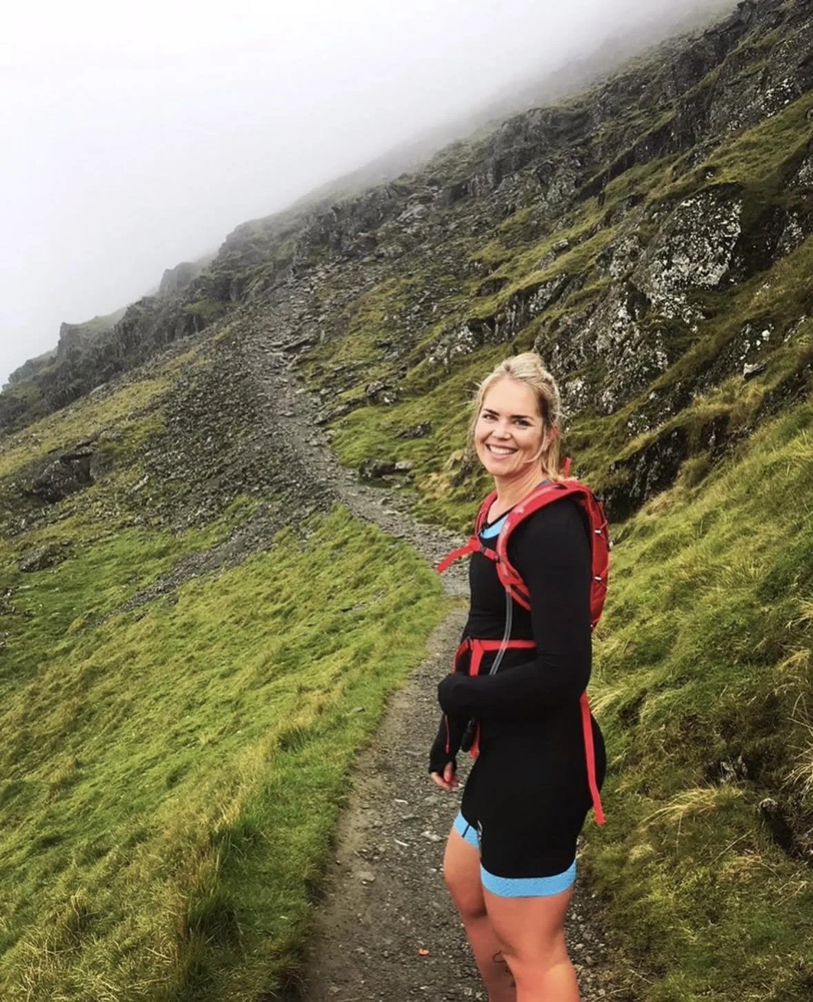 Smiling woman wearing black athletic clothing with blue accents, a red backpack, and a long sleeve shirt, standing on a narrow mountain trail surrounded by green grass and rocks, with fog-covered hillside in the background.
