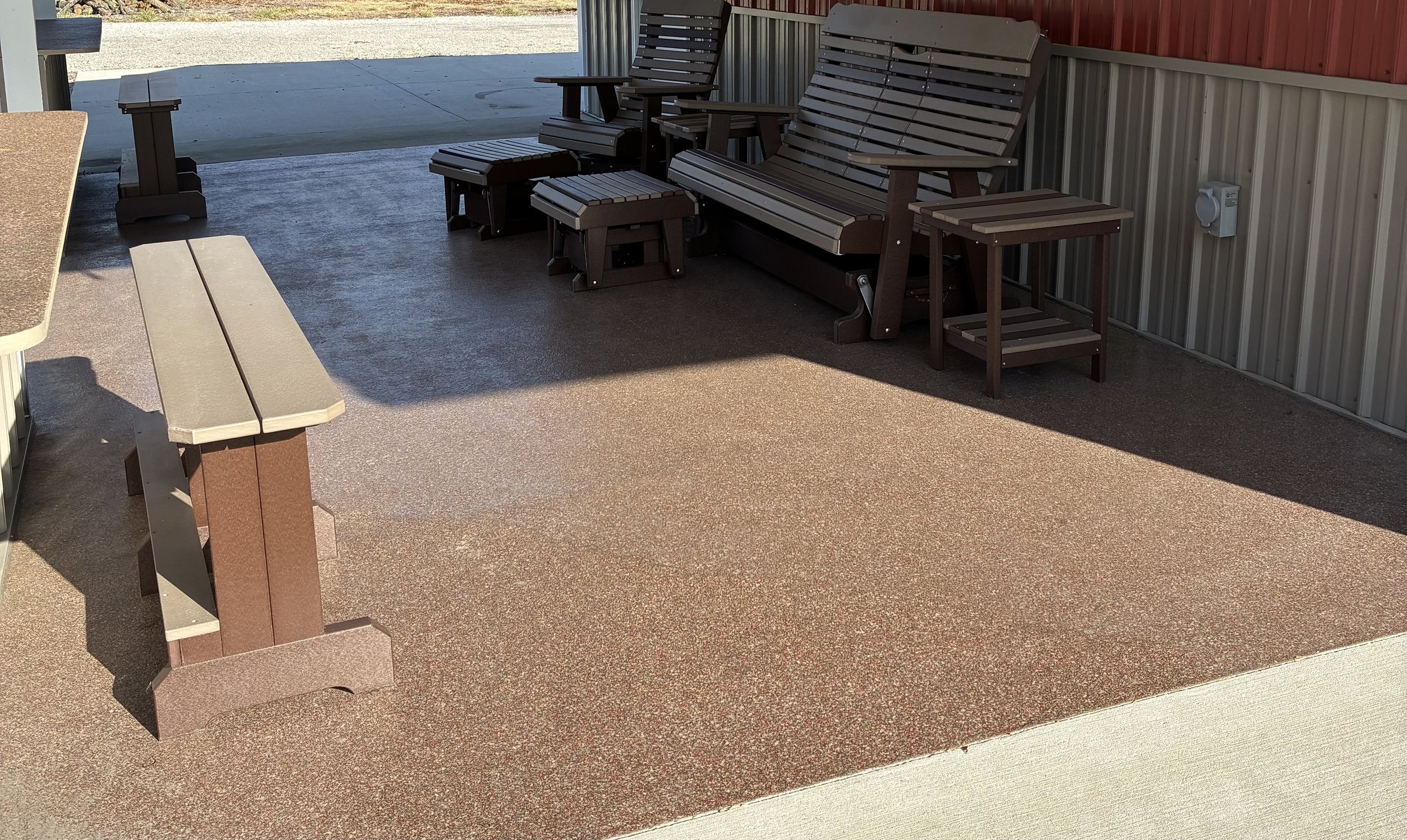 Outdoor seating area with benches, chairs, and tables on a textured ground, next to a building with metal siding.