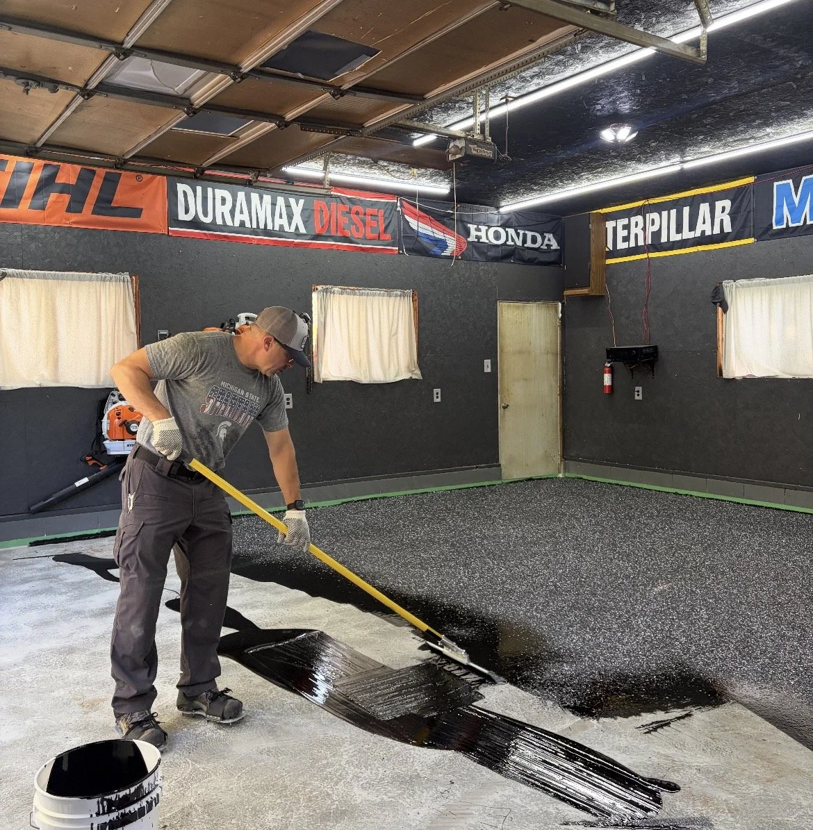 Man applying black waterproof coating on the floor of a garage with racing banner signs on the walls.