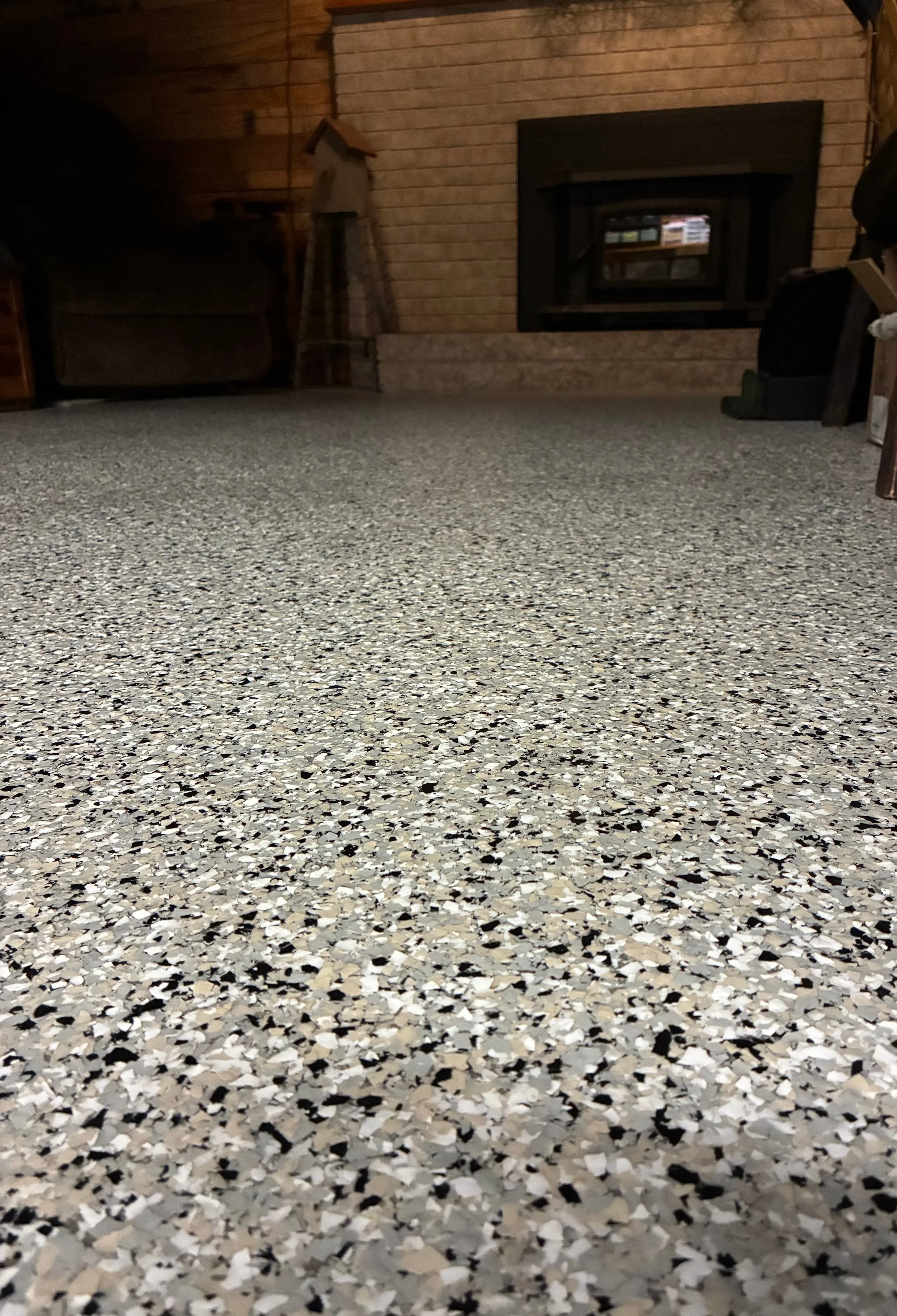 Close-up view of a pebble-textured carpeted floor in a basement with a brick fireplace and wood-paneled walls in the background.
