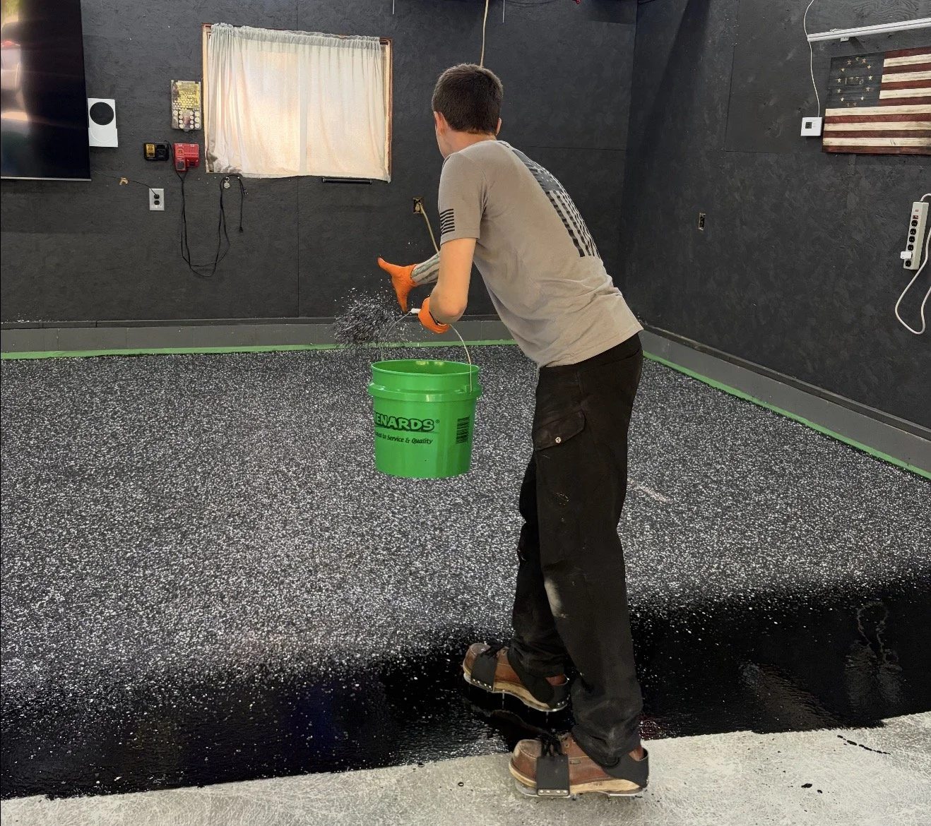 A man is spreading black epoxy resin on a garage floor. He is wearing gloves and work pants. The room has dark walls, a small window with a curtain, and various electrical outlets and tools on the walls. An American flag decoration is visible.