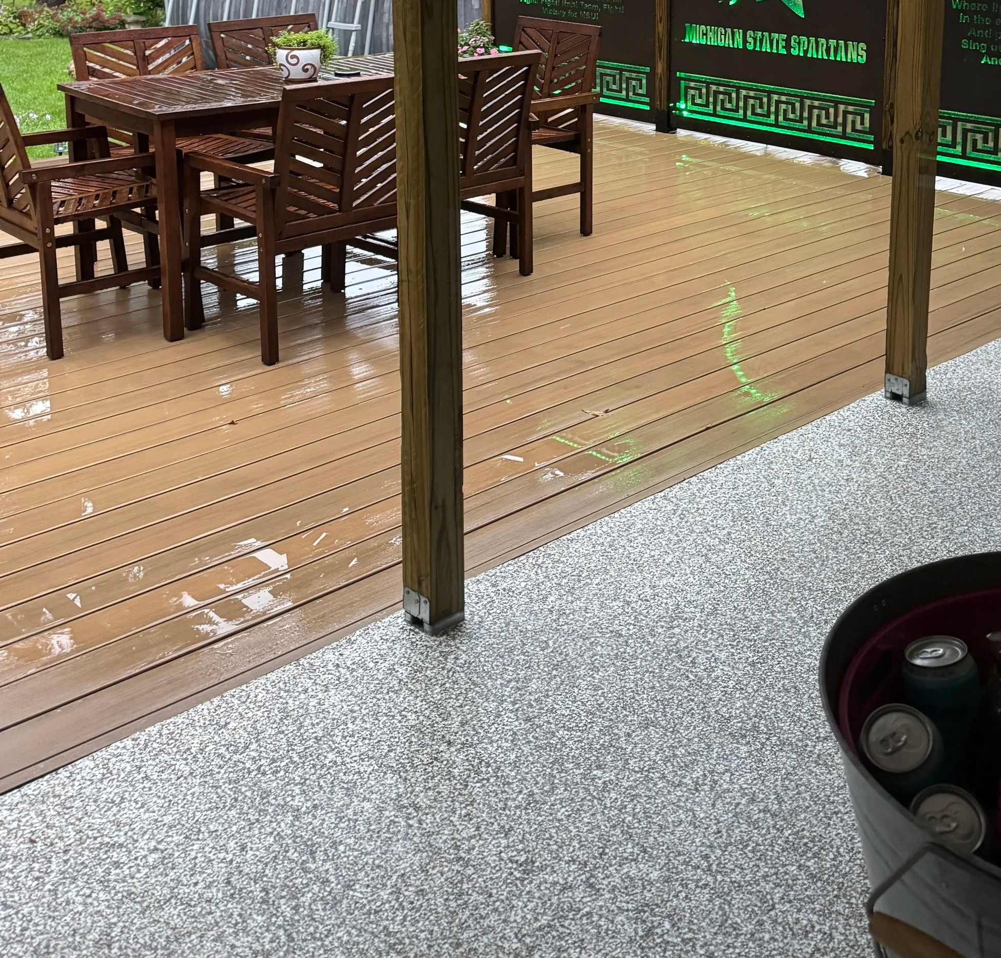 An outdoor patio with a brown wooden table and six matching chairs, wet from rain. A white pot with purple flowers is on the table. The patio has a wooden railing and a black sign with green text reading 'Michigan State Spartans.'