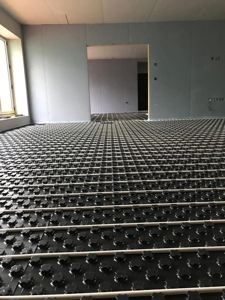 Interior of a building under construction with a radiant floor heating system installed, showing black panels with white pipes routed through them.