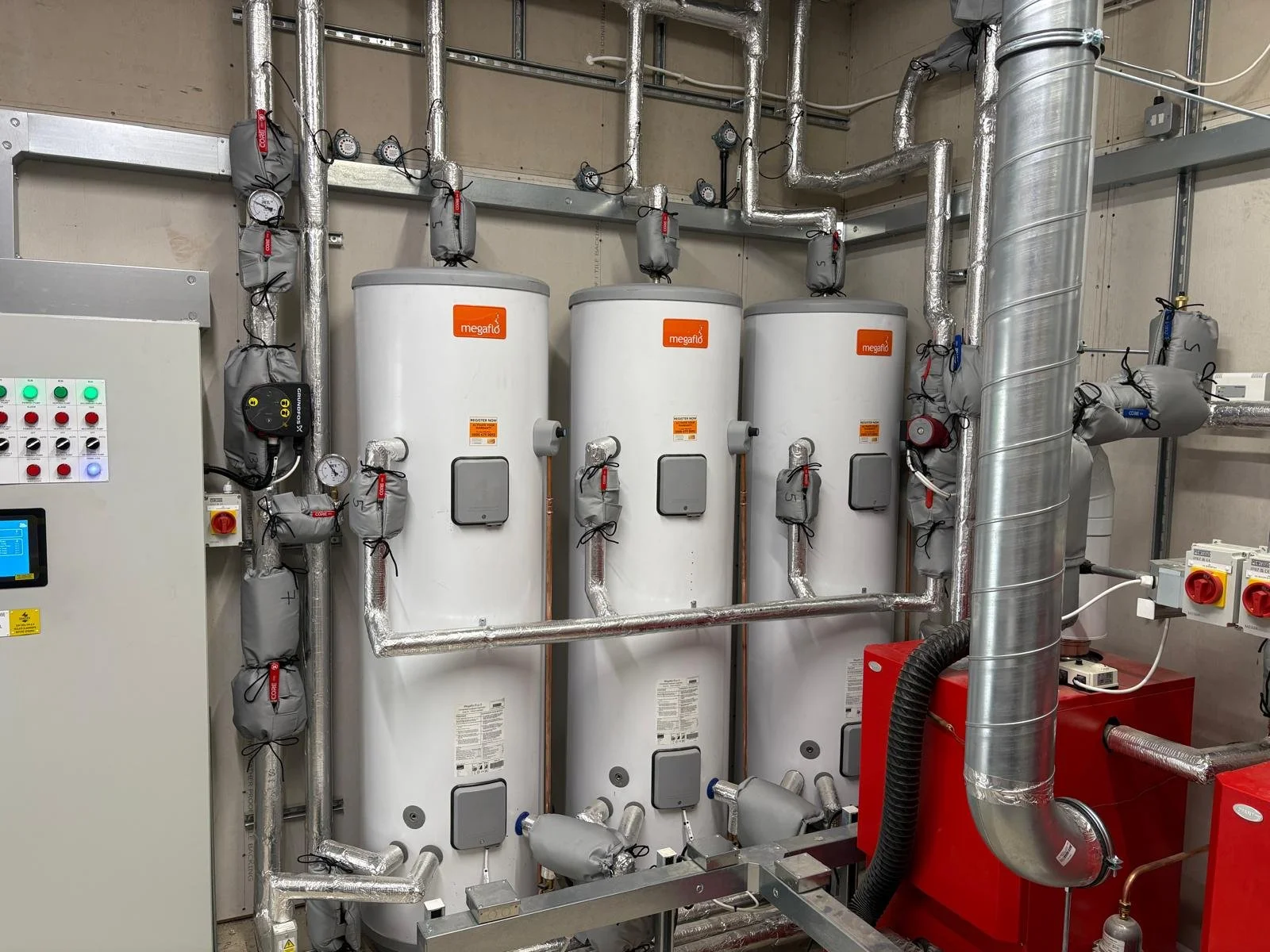 Three white vertical water heaters with the brand label 'megaflo' connected by pipes and surrounded by insulation wraps, with control panels, gauges, and additional industrial equipment in a mechanical room.
