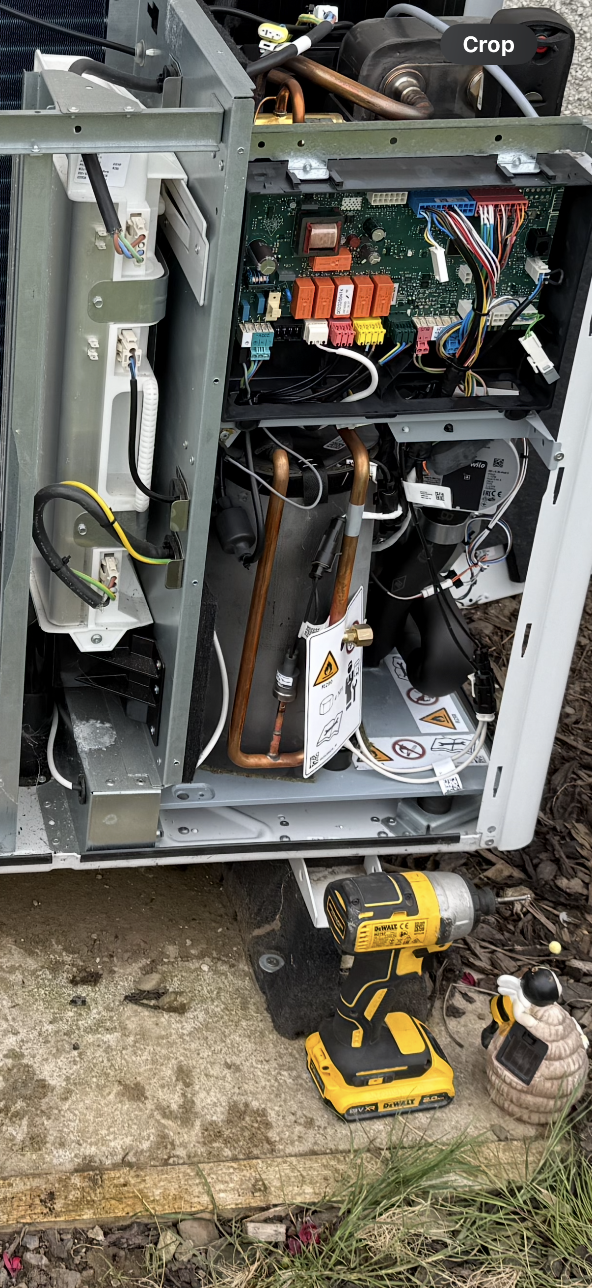 Open HVAC unit showing internal components, copper pipes, circuit boards, wiring, and a yellow cordless drill on the ground outside the unit.