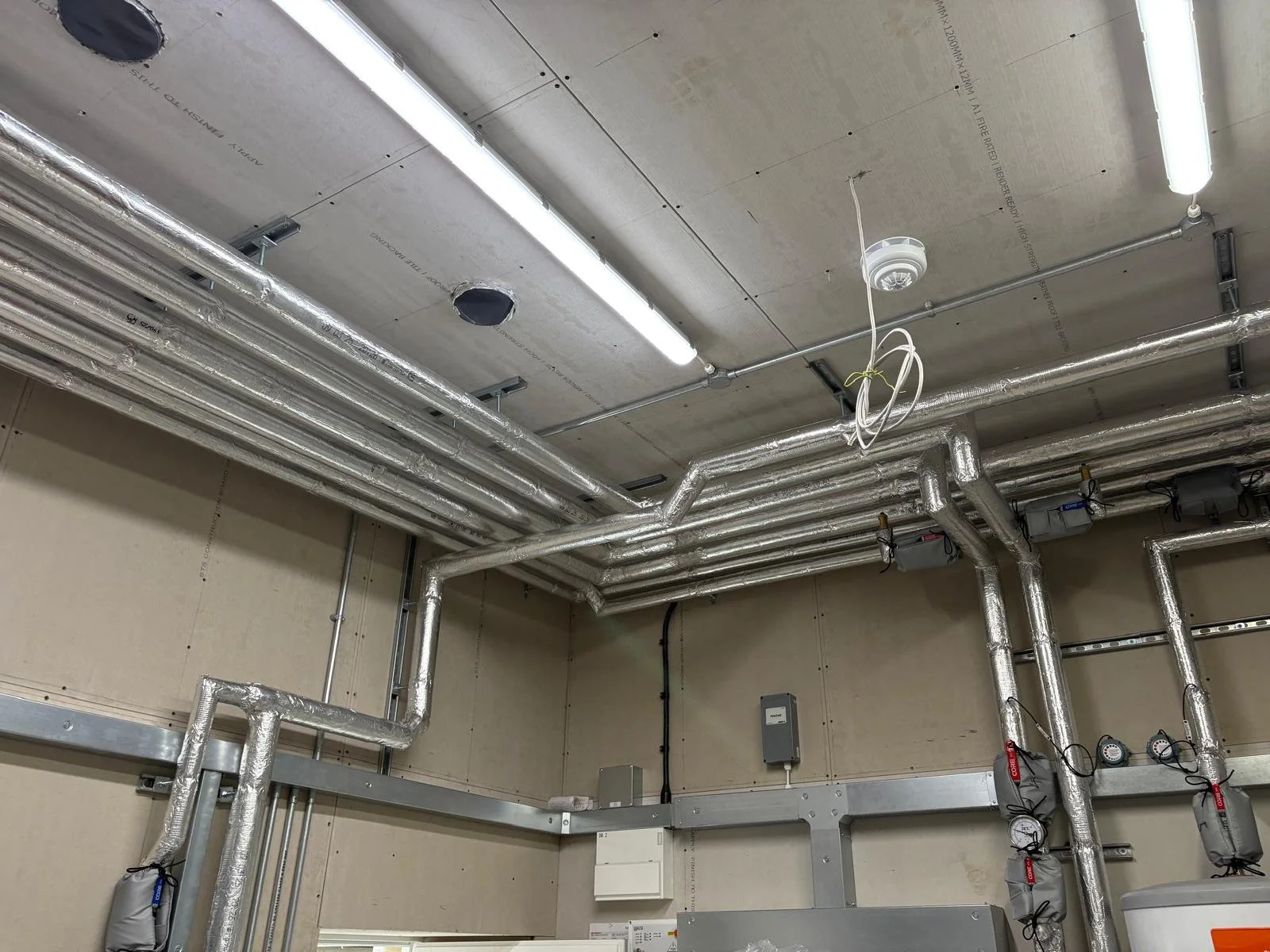 A construction site ceiling with exposed ductwork wrapped in silver insulation, electrical wiring, and fluorescent light fixtures.