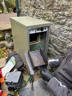 An old, empty metal vending machine outdoors, with the front panel removed, exposing its interior components and wiring, next to a stone wall.
