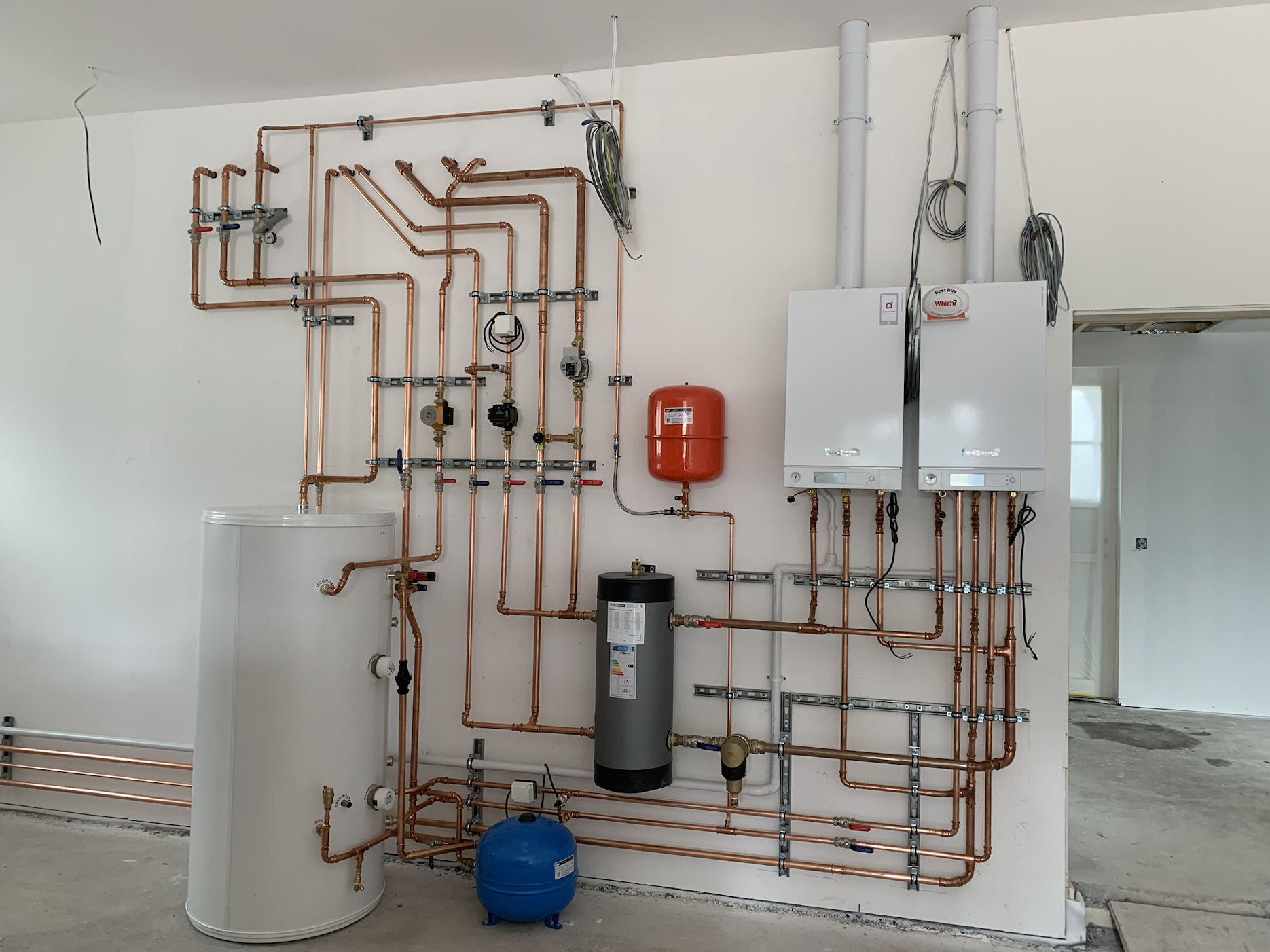 Interior view of a plumbing installation with copper pipes, valves, water heater, expansion tank, filter, and other components on a white wall in a building under construction or renovation.