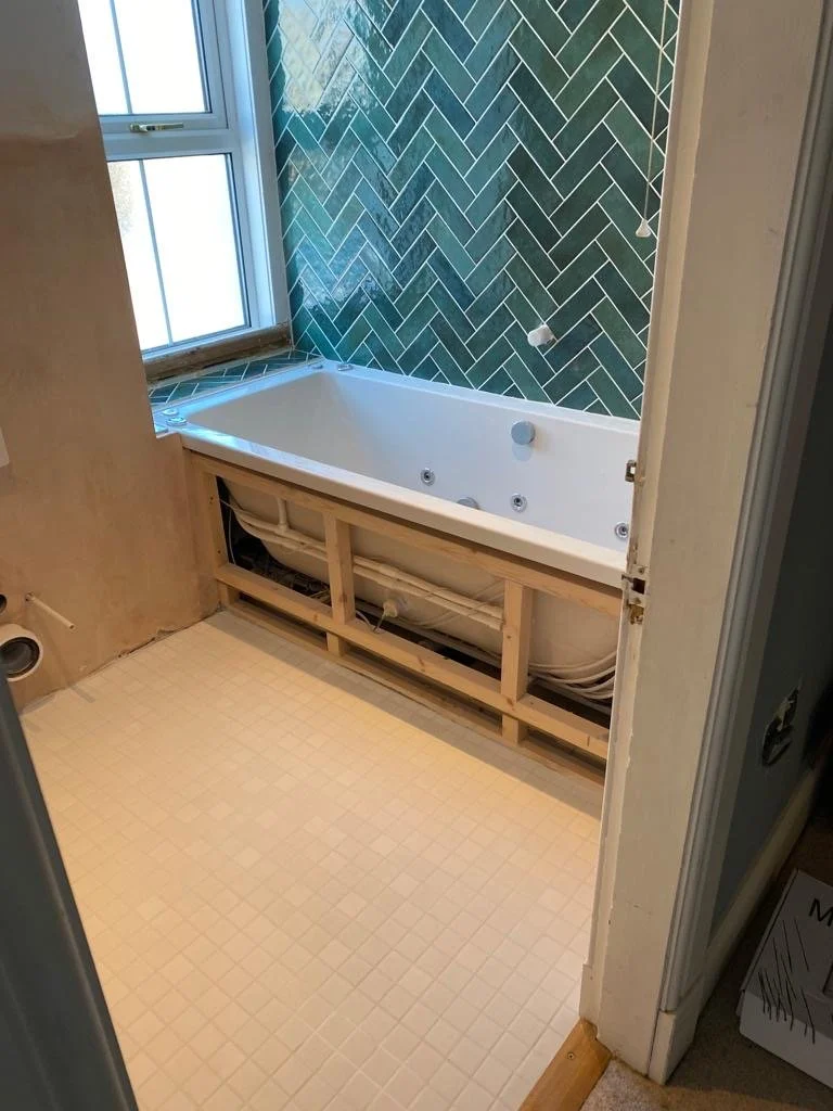 Bathroom with a partially installed soaking tub, green herringbone tile on the wall, and a window. The floor has small beige tiles, and the area below the tub is unfinished with visible pipes and framing.