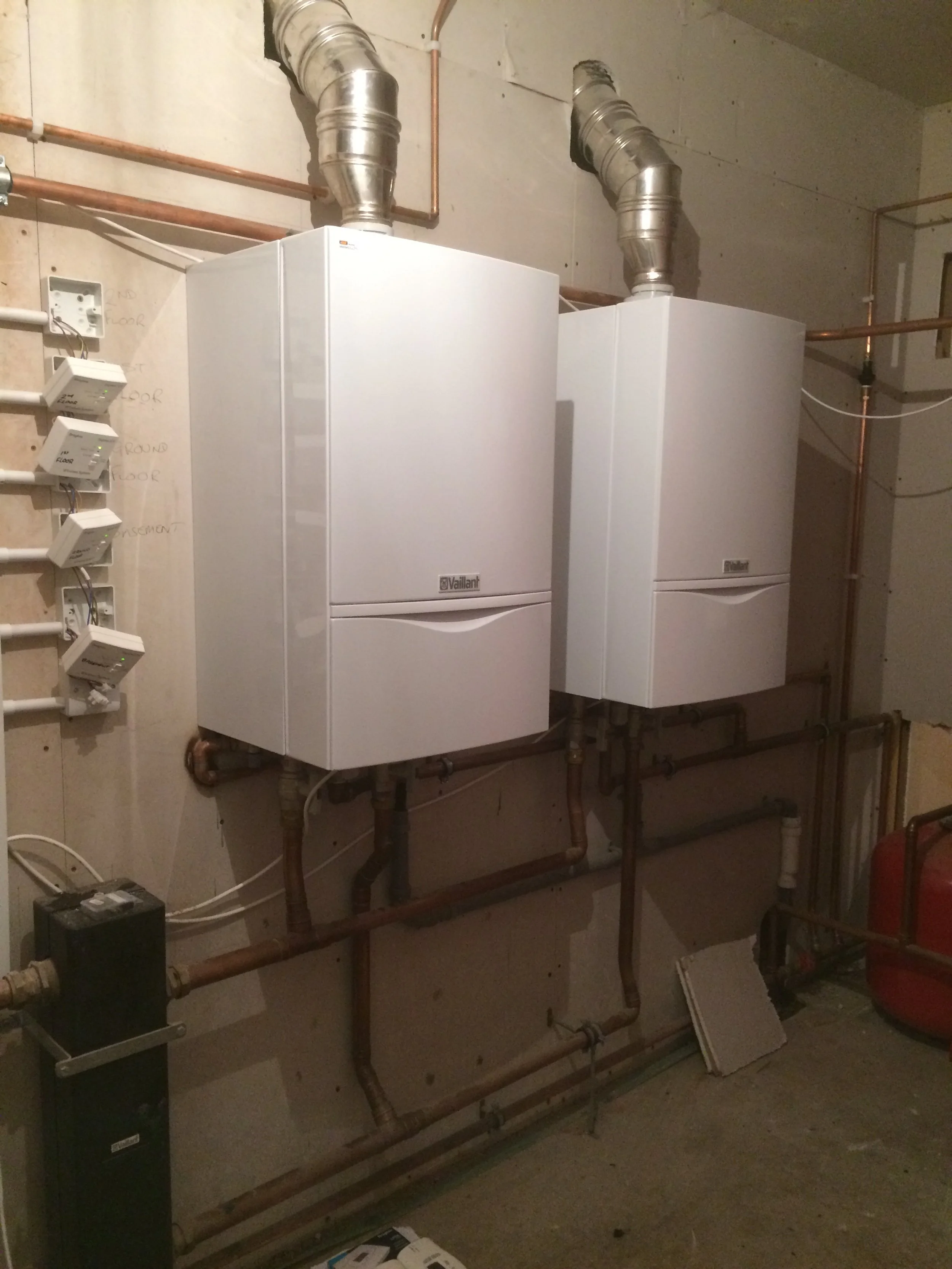 Two white Vaillant boiler units mounted on a wall in a utility room with copper and PVC pipes, a red expansion tank, and electrical components.