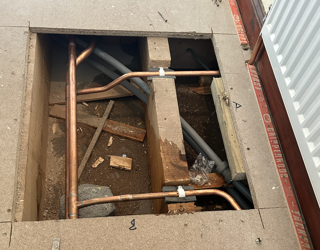 Open floor with exposed plumbing pipes and electrical wiring, surrounded by construction debris and framing materials.