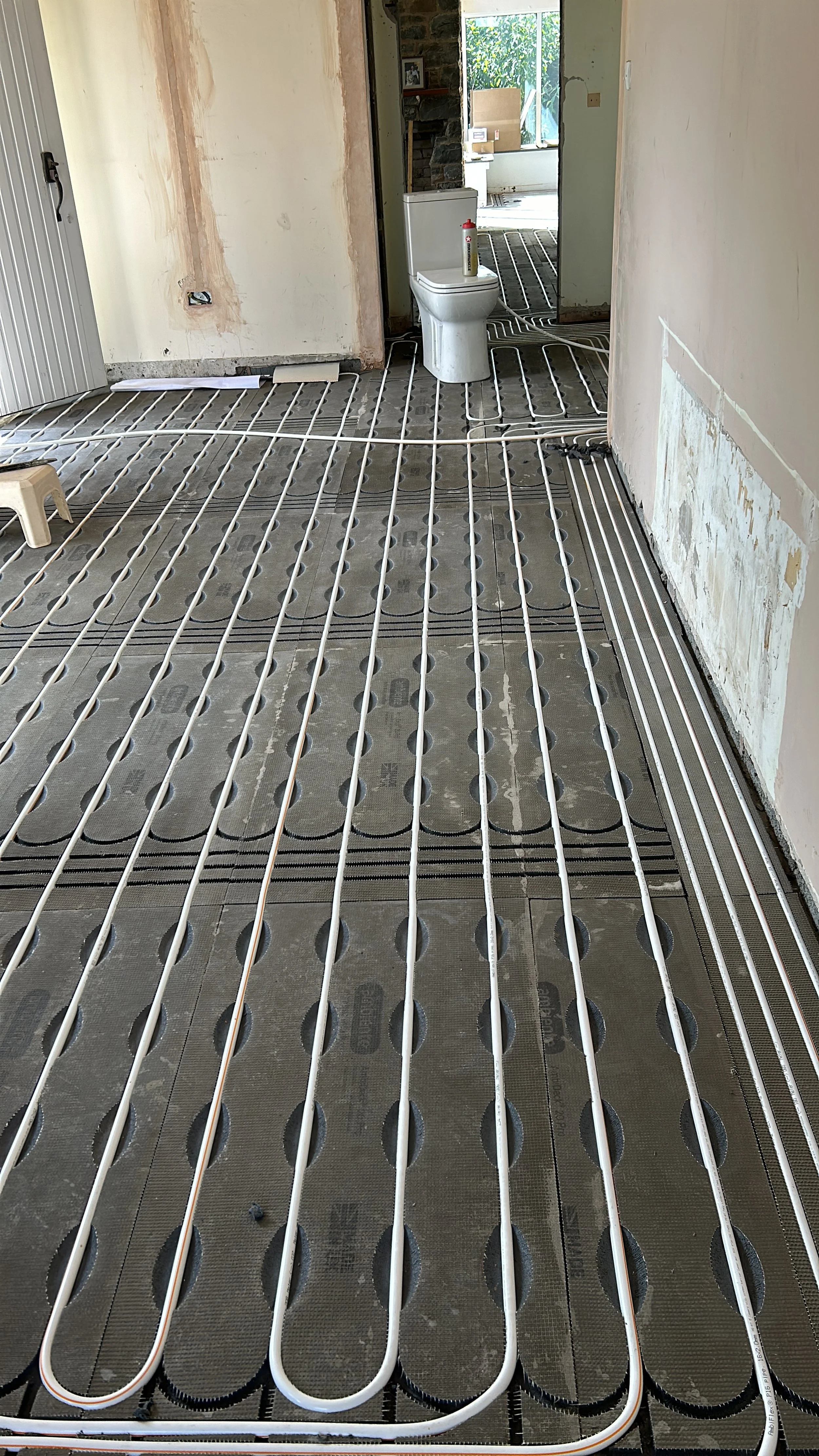 Underfloor heating pipes installed in a room, with a toilet and a doorway leading to another room in the background.