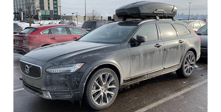 Voiture de police noire avec un coffre de rangement sur le toit, stationnée dans un parking. La voiture est sale et le parking est rempli d'autres véhicules.