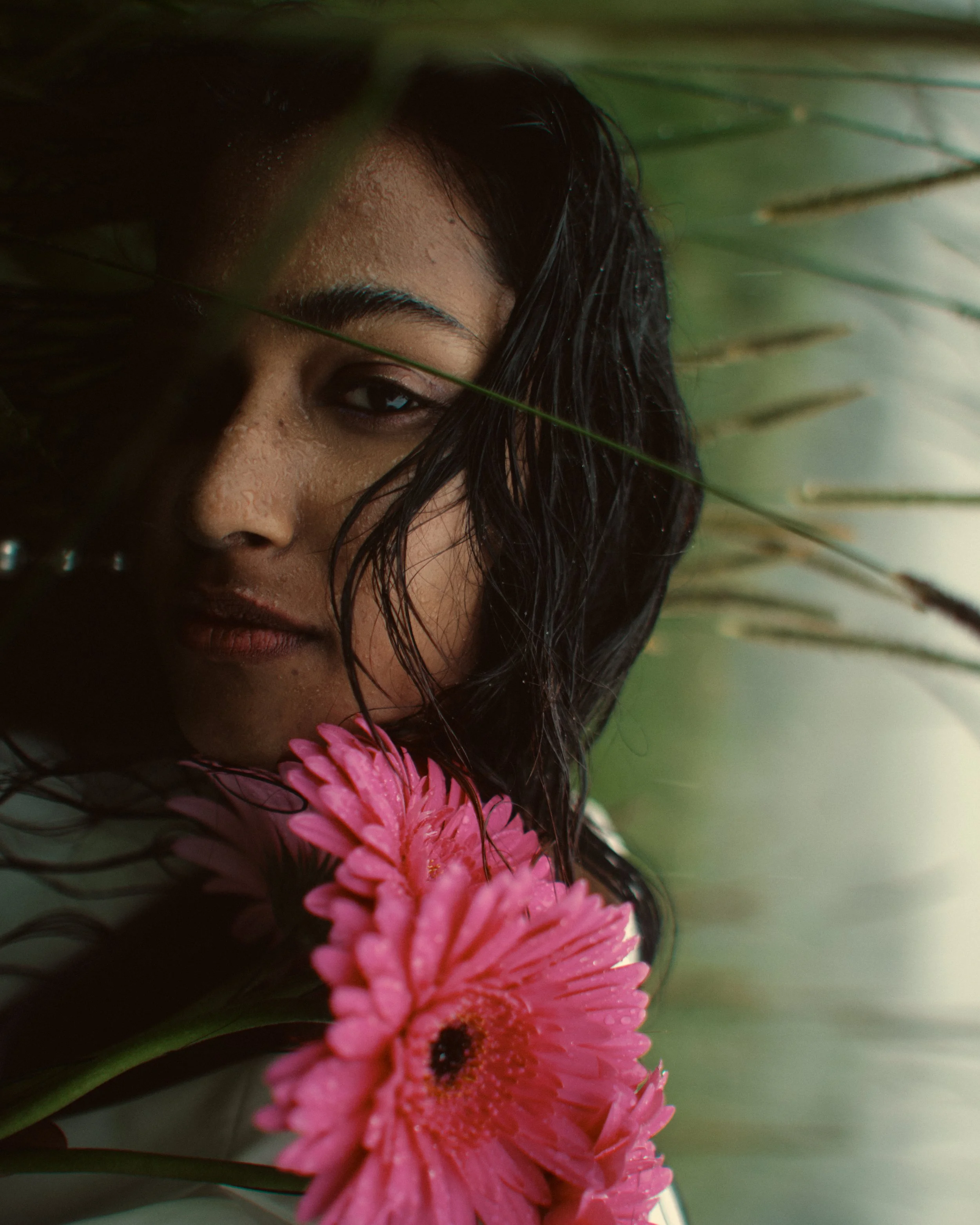 A woman with wet hair holds a pink flower near her face, partially obscured by green leaves.