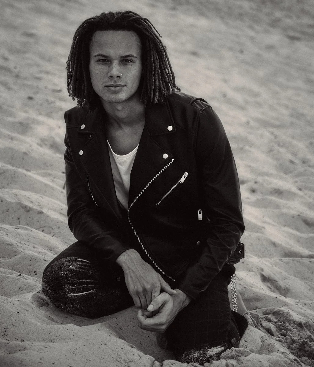 Black and white photo of a young man with dreadlocks, wearing a leather jacket and white shirt, sitting on sand with one knee up and hands clasped.