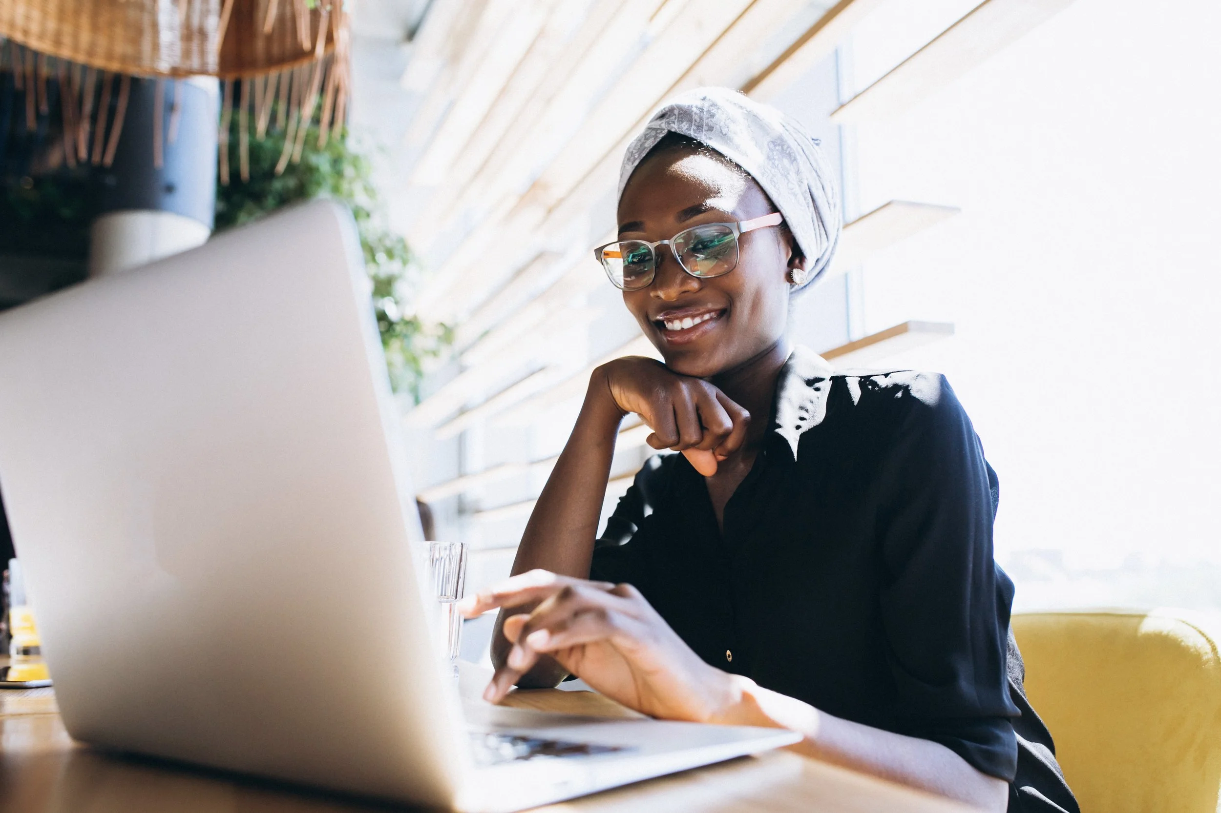 A woman with glasses and a headscarf smiling at her laptop in a bright, modern space.