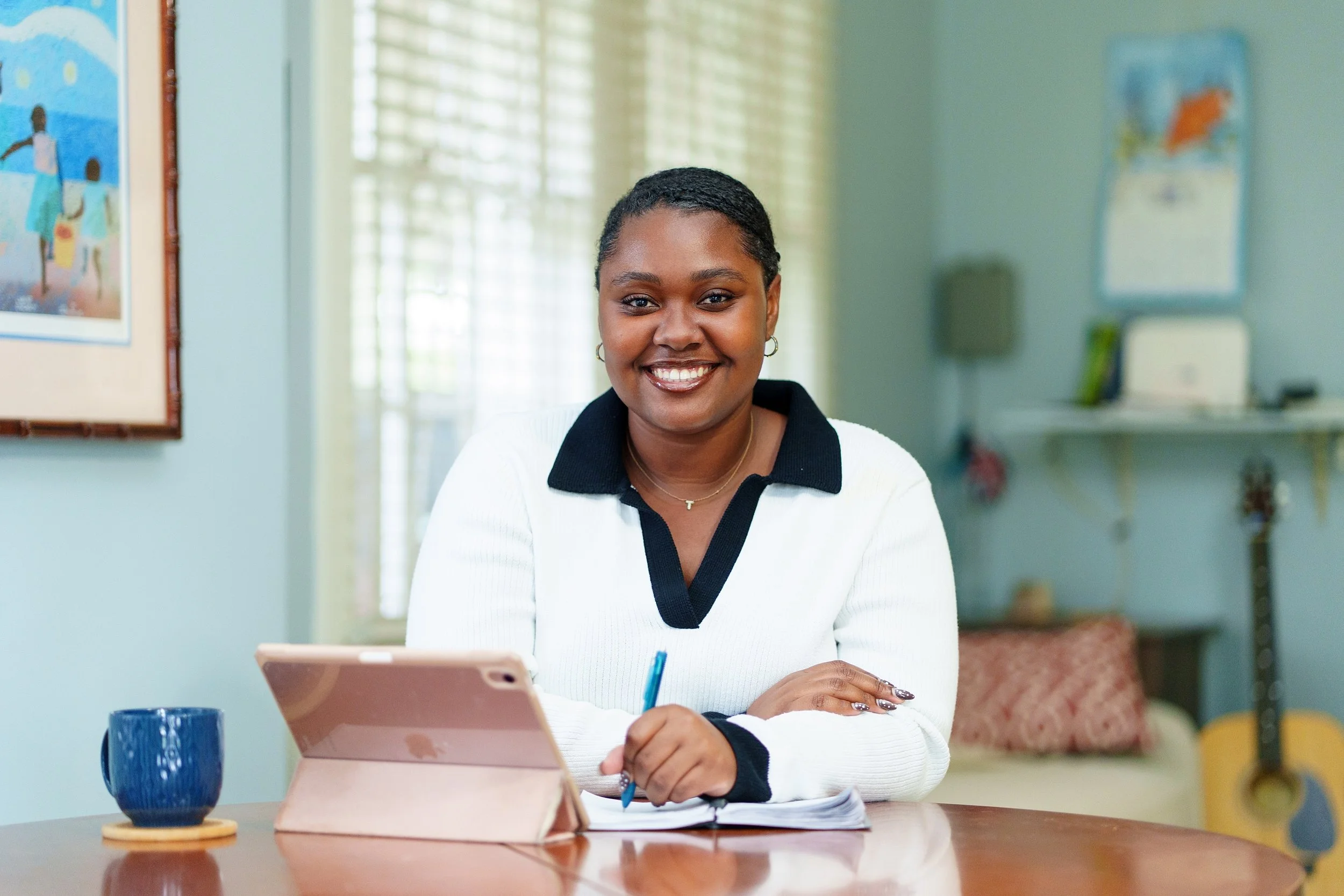 A woman sitting at a wooden table with a tablet, a cup, and some papers, smiling at the camera in a cozy room with artwork and a guitar in the background.