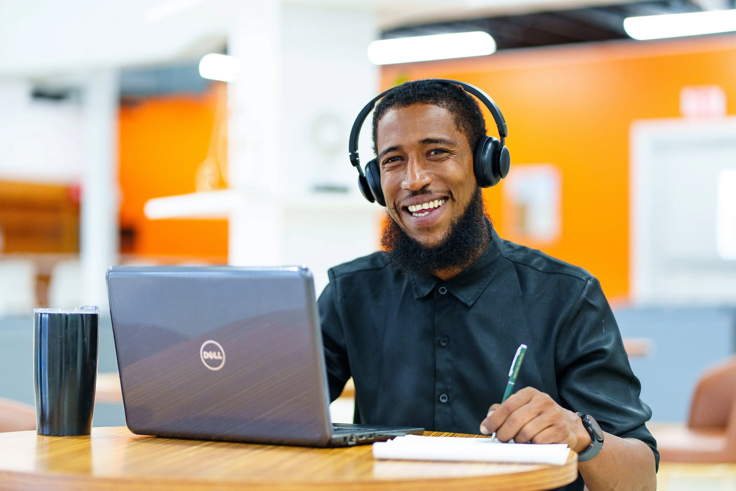A smiling man wearing headphones sitting at a desk with a laptop, pen, and notebook in a modern office environment.
