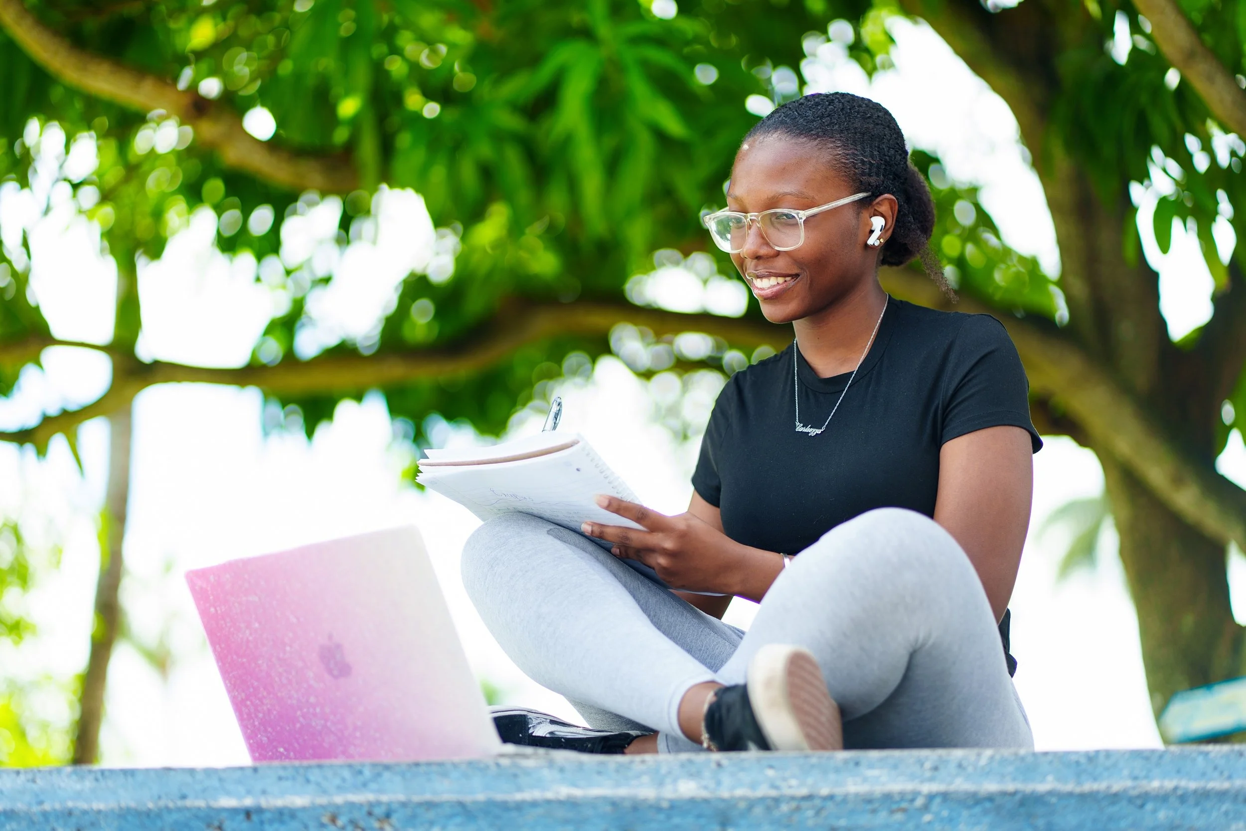 Woman outdoors sitting on a blue surface with a pink laptop, holding a notebook and writing, surrounded by green trees.